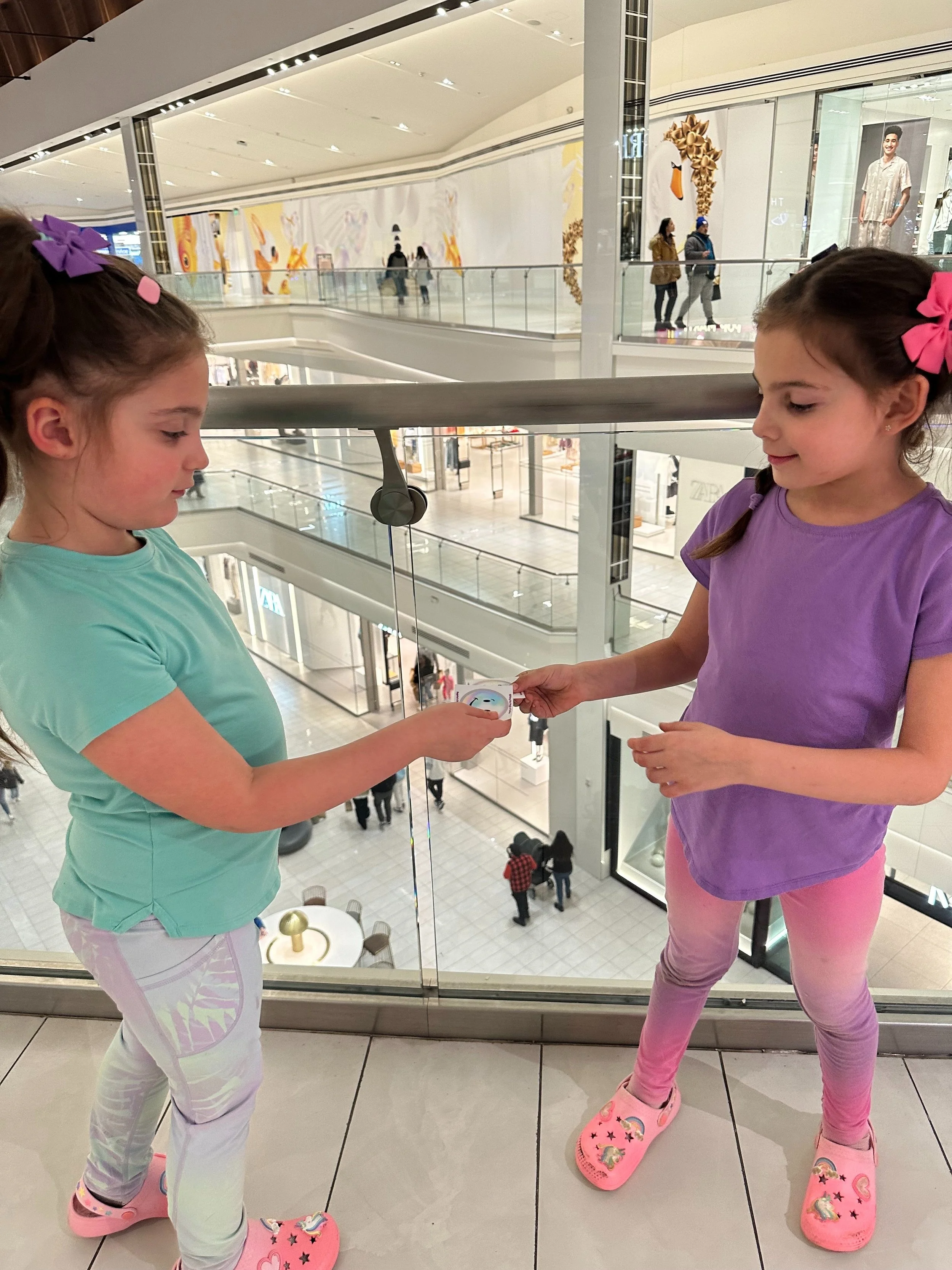 Two young girls standing on an upper level of a shopping mall, exchanging a small object. The girl on the left wears a light blue top and pastel patterned pants, while the girl on the right wears a purple shirt and pink leggings. They are both wearin