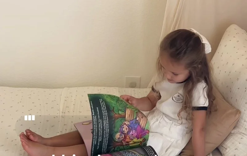 Young girl sitting on a bed, reading a colorful illustrated book.