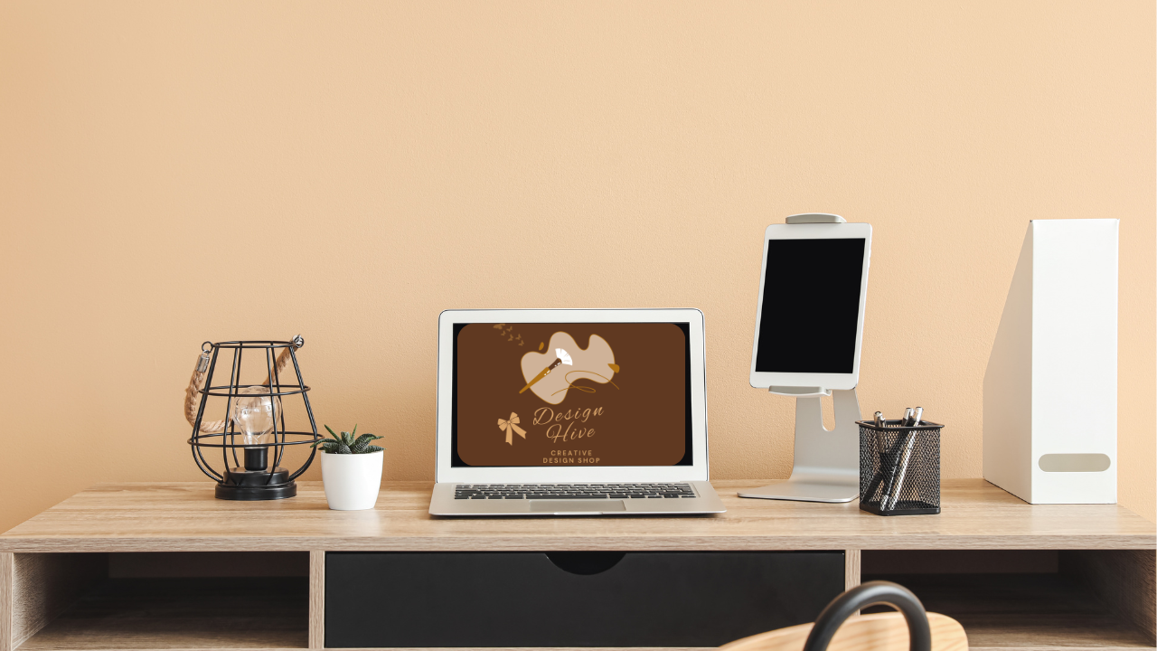 Office desk with computer, laptop, phone stand, illuminated wireframe lamp, small potted plant, pen holder, and storage box against a beige wall.