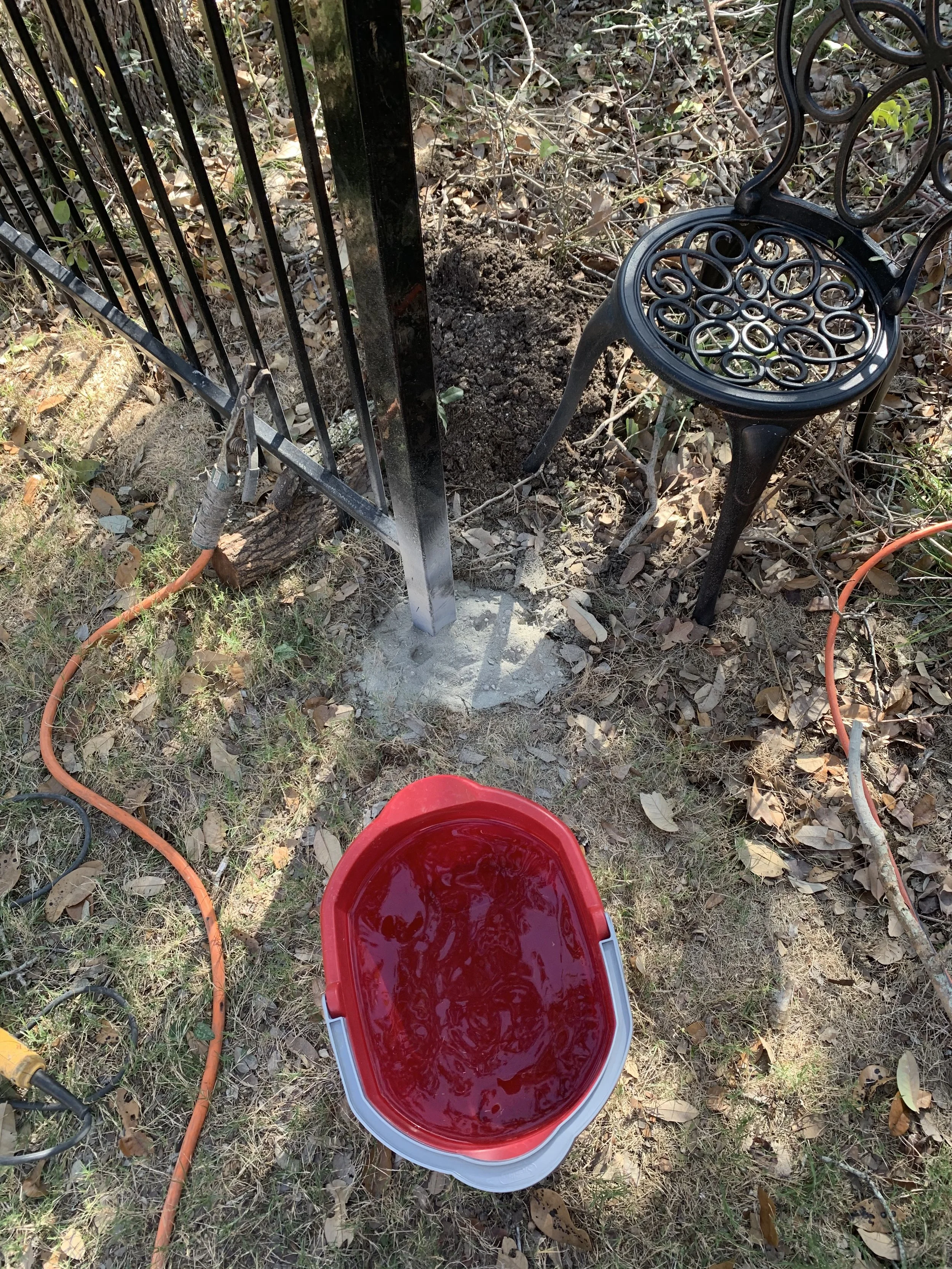 A garden scene with a red bucket filled with water, a black metal chair, and a black metal fence, with dirt, leaves, and some plants around.