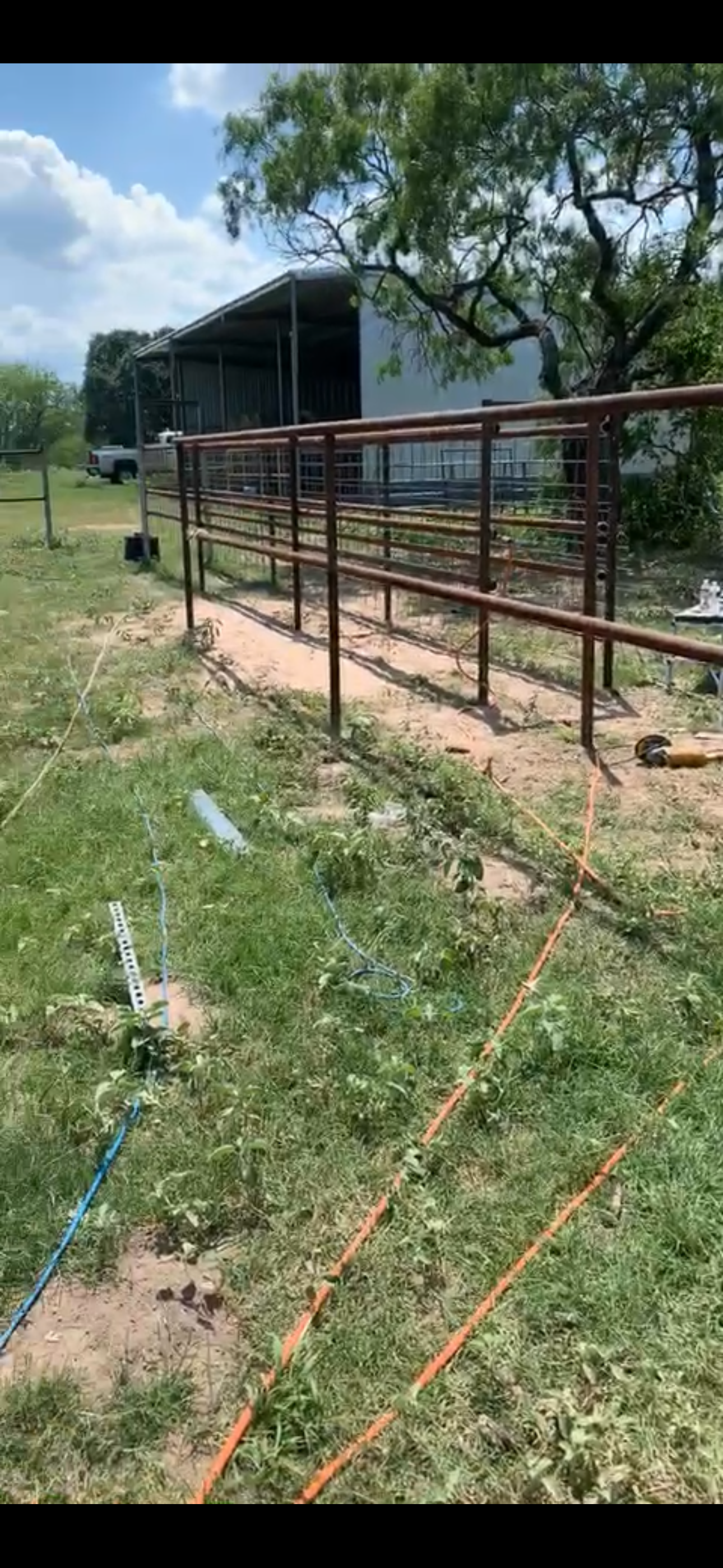 Construction site with metal framework, electrical wiring on the ground, and a large tree under a partly cloudy sky.