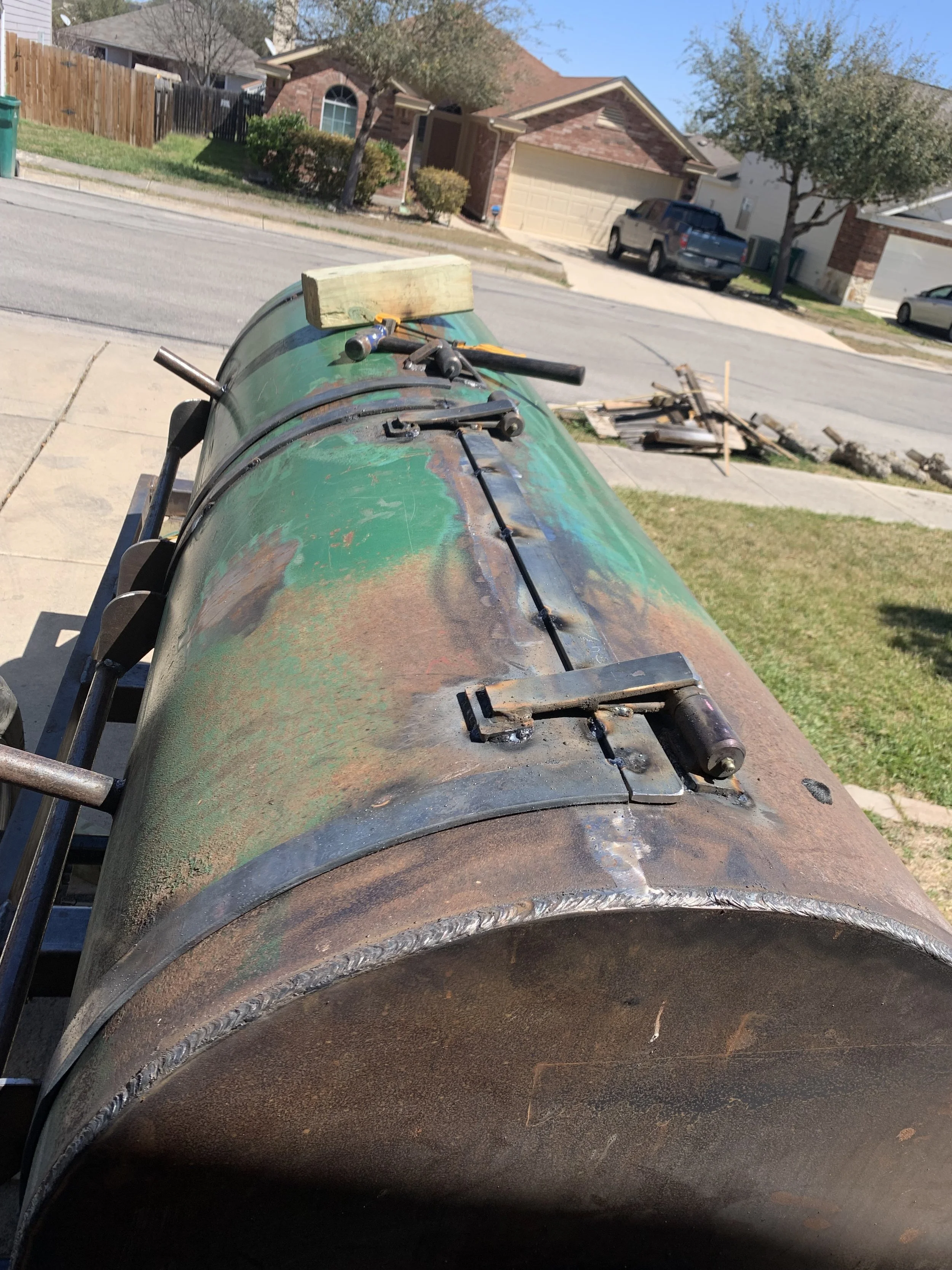 A large metal cylindrical object, possibly an industrial or construction piece, with tools placed on top, situated outdoors in a residential neighborhood with houses, trees, and parked cars visible in the background.