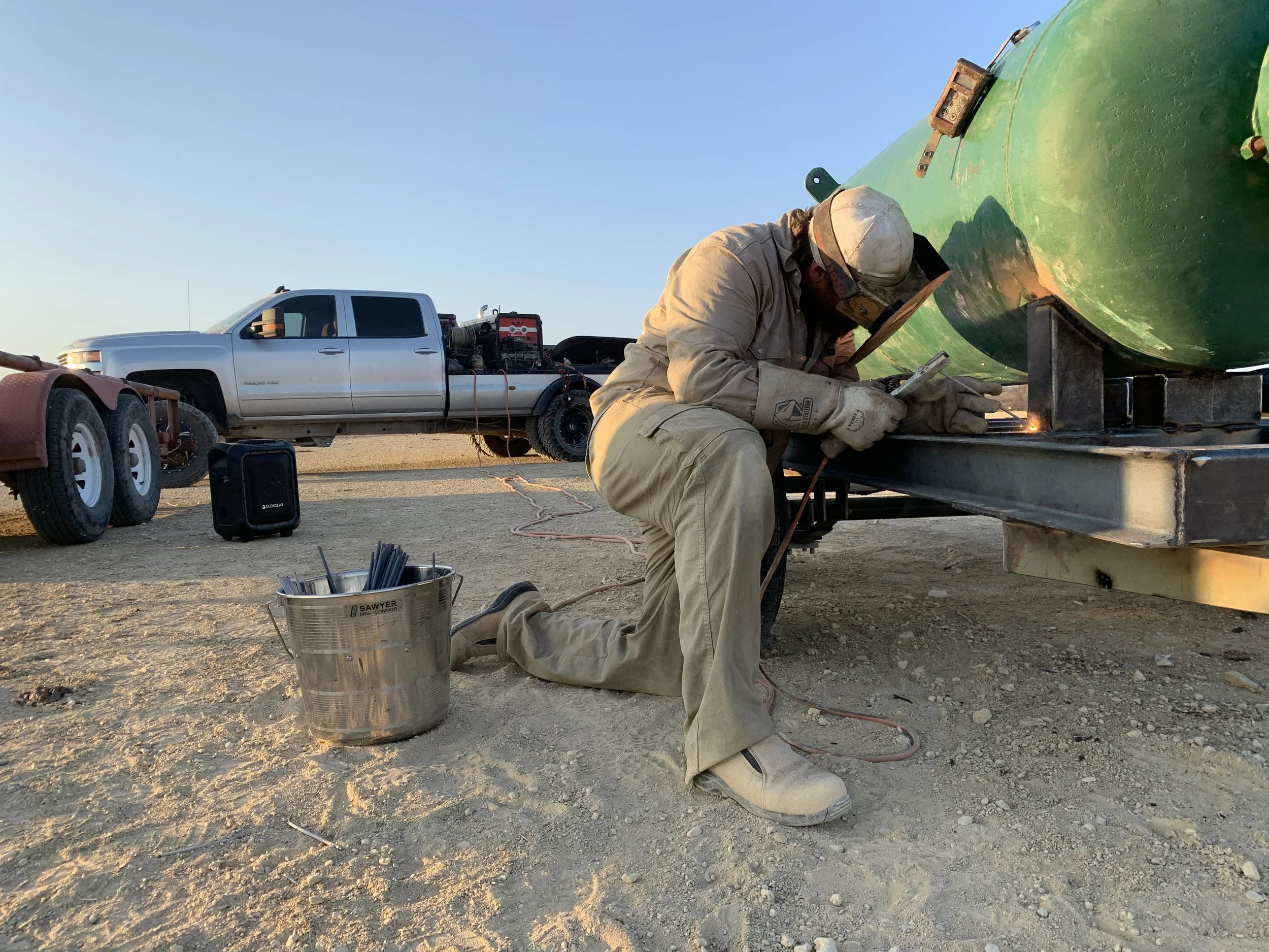 A person welding on a large green boat hull outdoors during sunset. The person is kneeling and wearing protective gear, including a helmet, gloves, and a jacket. Nearby are a bucket filled with welding rods, a small black speaker, and a white pickup truck with equipment in the bed, parked on a dirt surface.