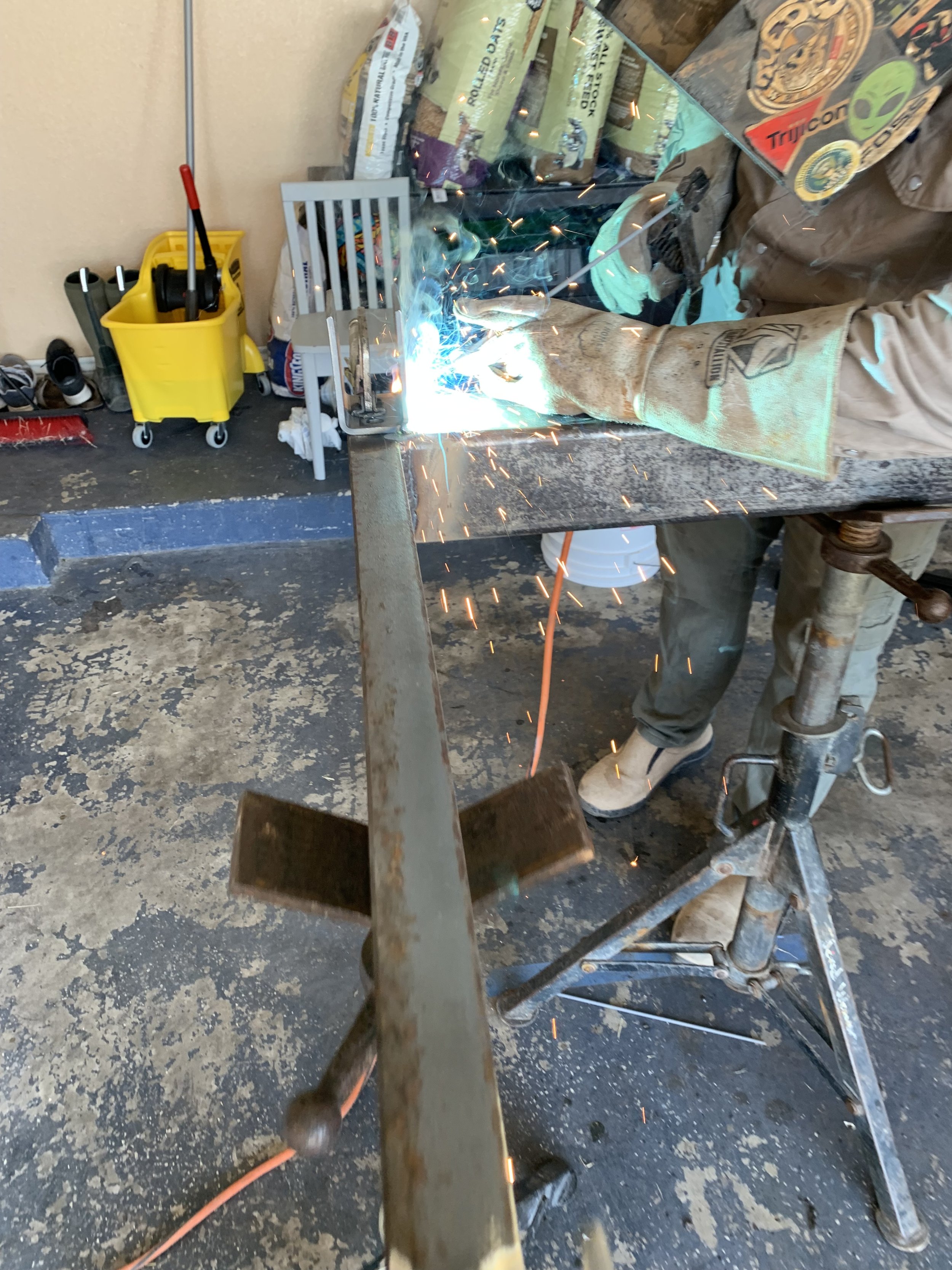Person welding a metal piece in a workshop with sparks flying, wearing protective gloves and gear.