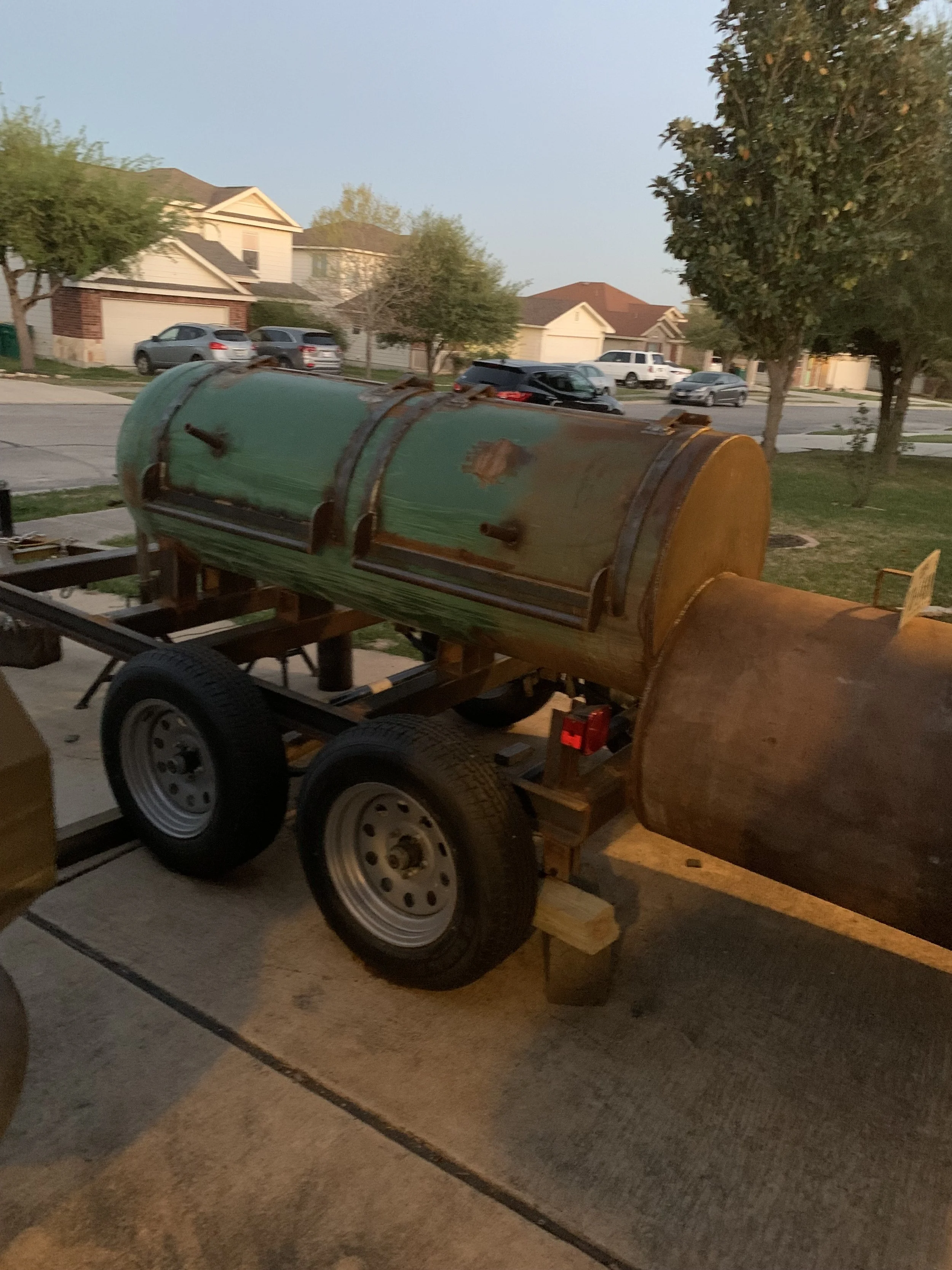 A rusty metal barbecue smoker on a trailer parked on a driveway with houses and trees in the background.