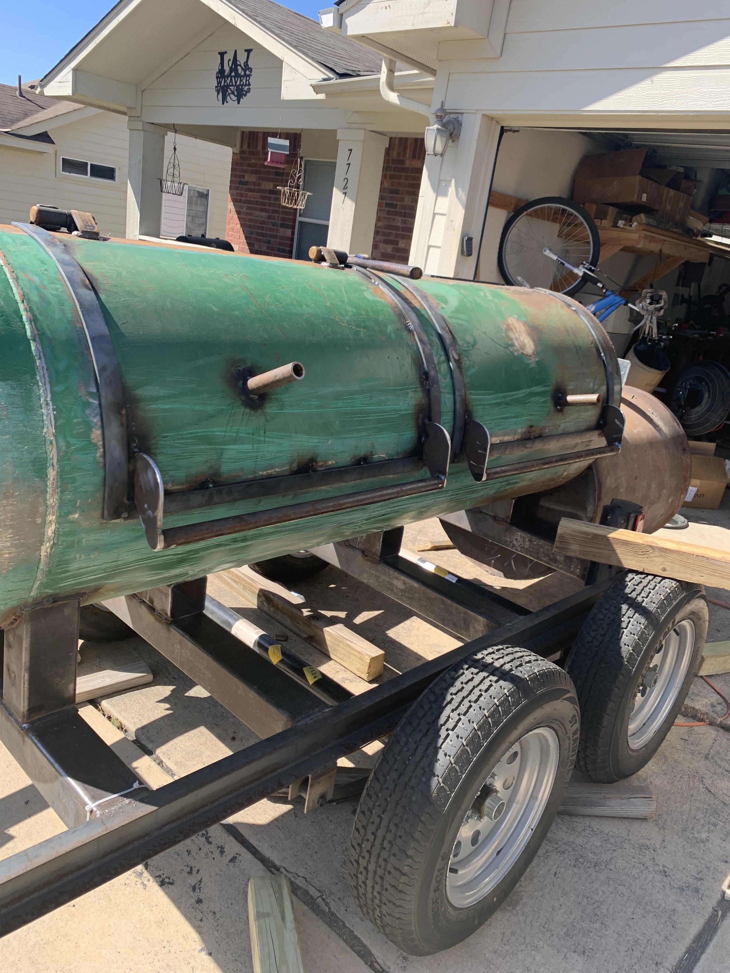 A large green metal tank on a wooden trailer with two black wheels, parked outdoors in front of a house with white siding, a brick chimney, and a garage with various tools and equipment inside.