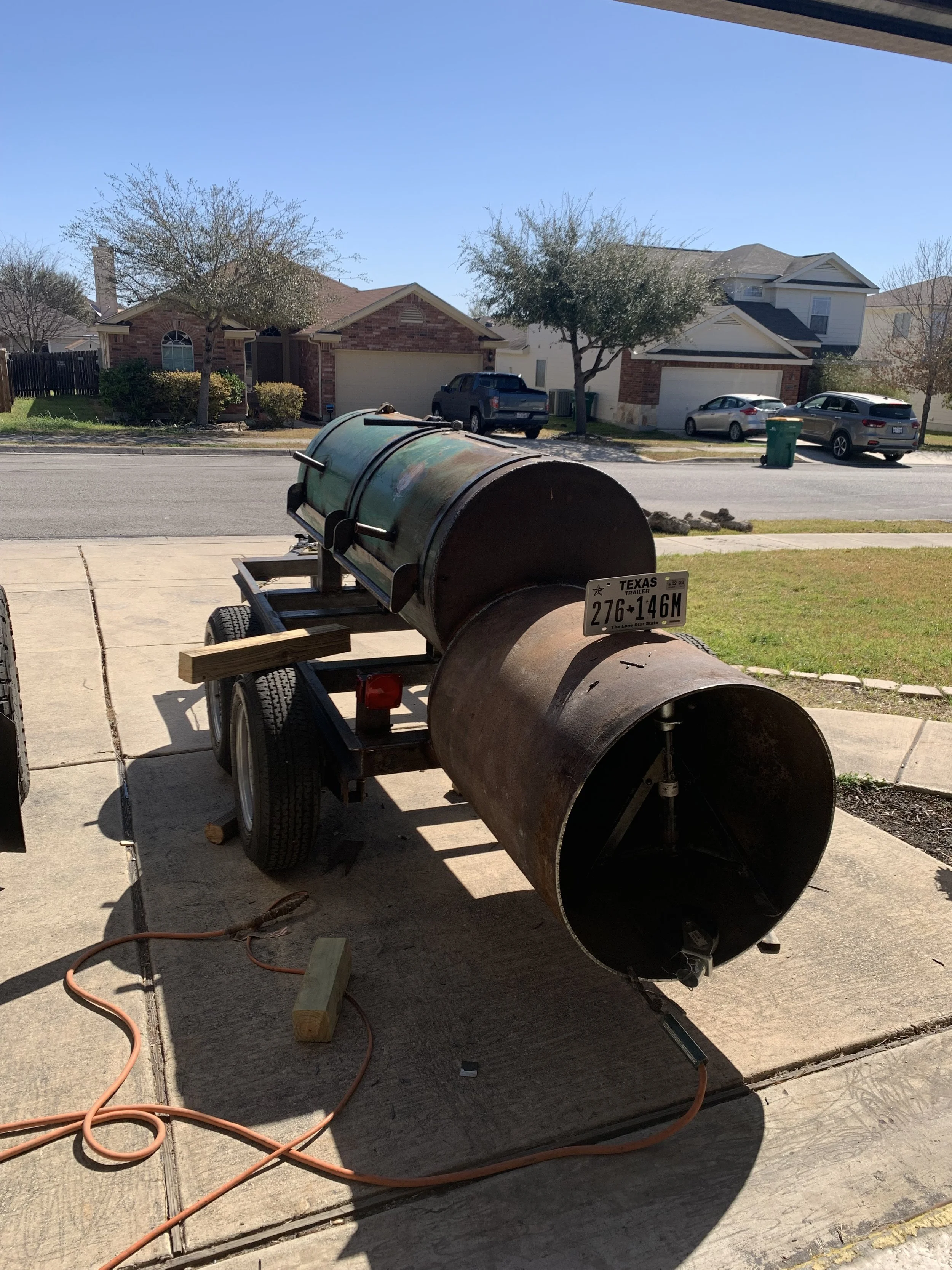 A rusty metal barbecue smoker on a trailer parked on a driveway with extension cords, wooden blocks, and small rocks, with houses and trees showing in the background.