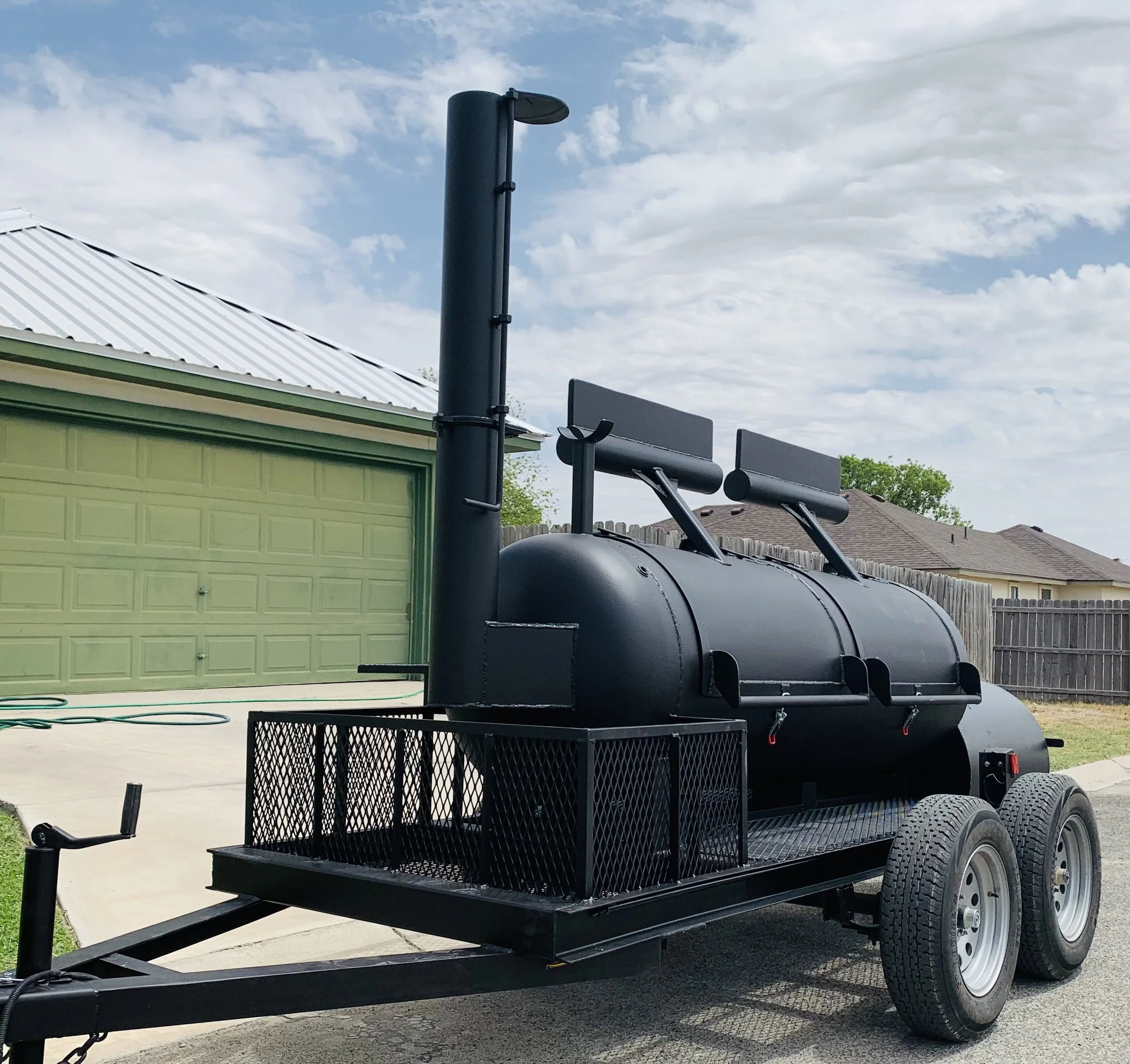 A black trailer with a barbecue smoker on it, parked in a driveway next to a green garage and a wooden fence, with houses and trees in the background and a blue sky with scattered clouds.