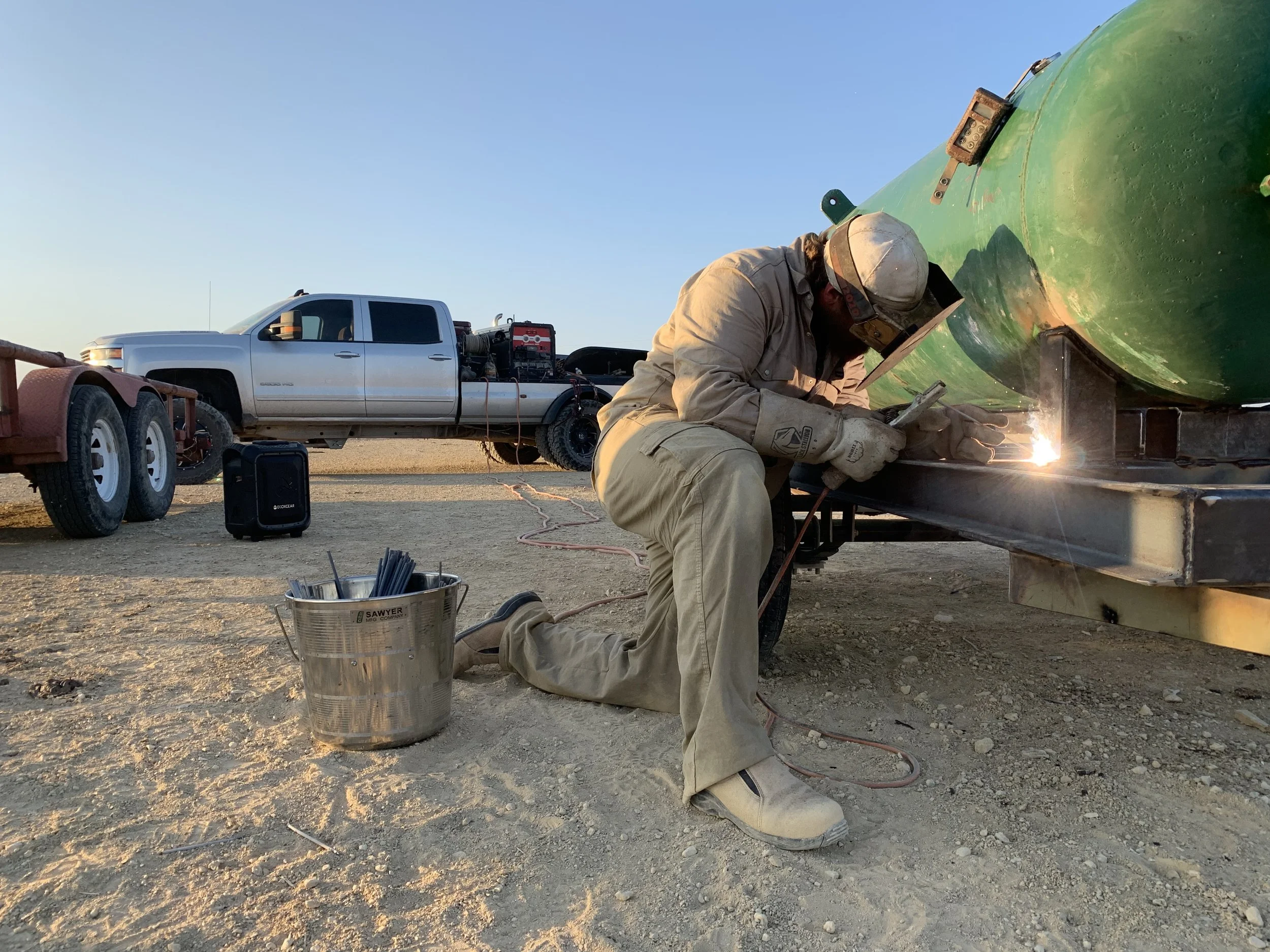 A person welding on a green boat in an outdoor setting with a white pickup truck in the background. The person is wearing beige work clothes and a welding helmet. Sparks from welding are visible. Various tools and equipment are nearby.