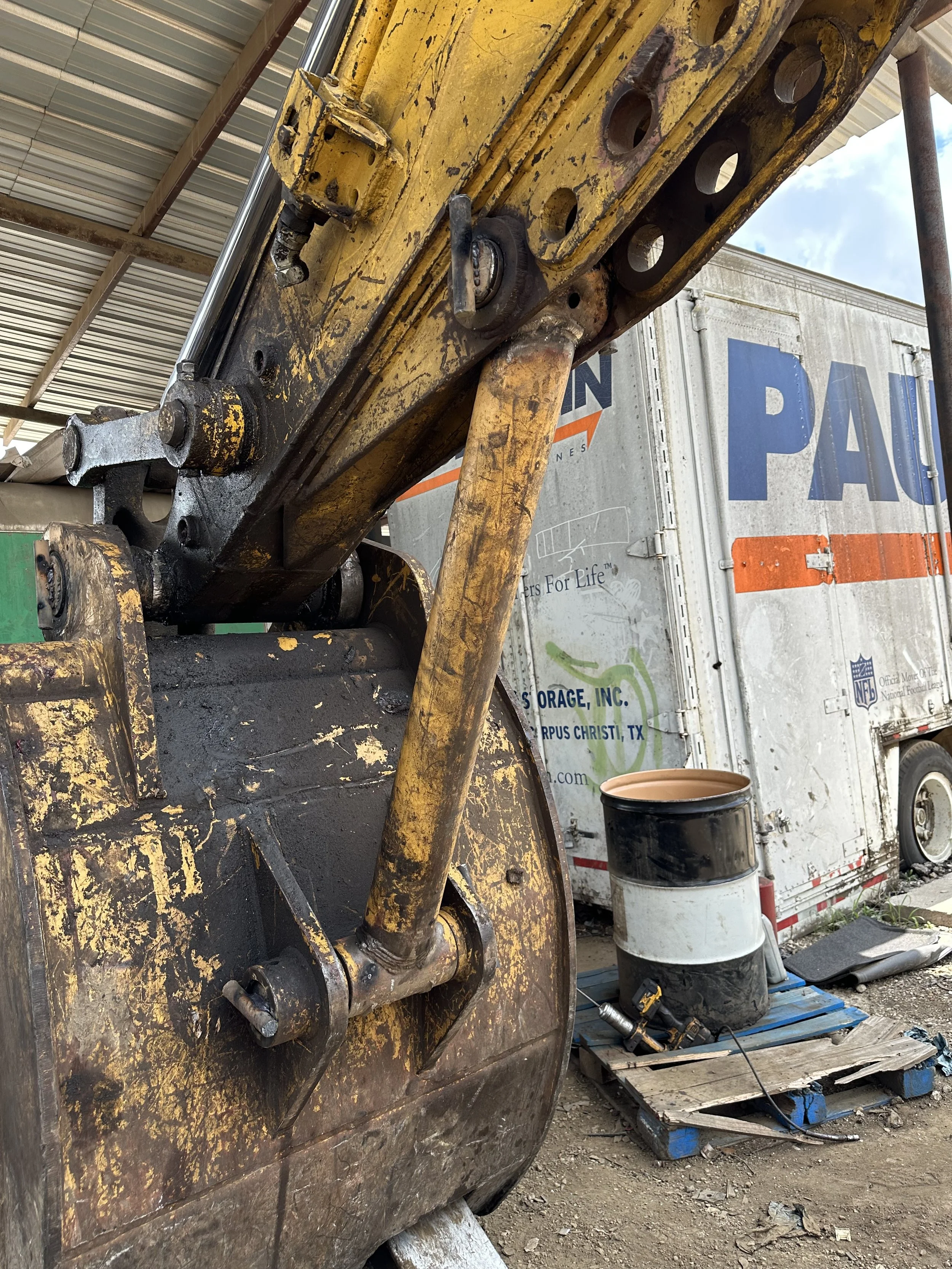 Close-up of a large, yellow, and black excavator arm with visible hydraulic cylinder and mechanical parts, parked outdoors near a trailer with a partially visible sign.