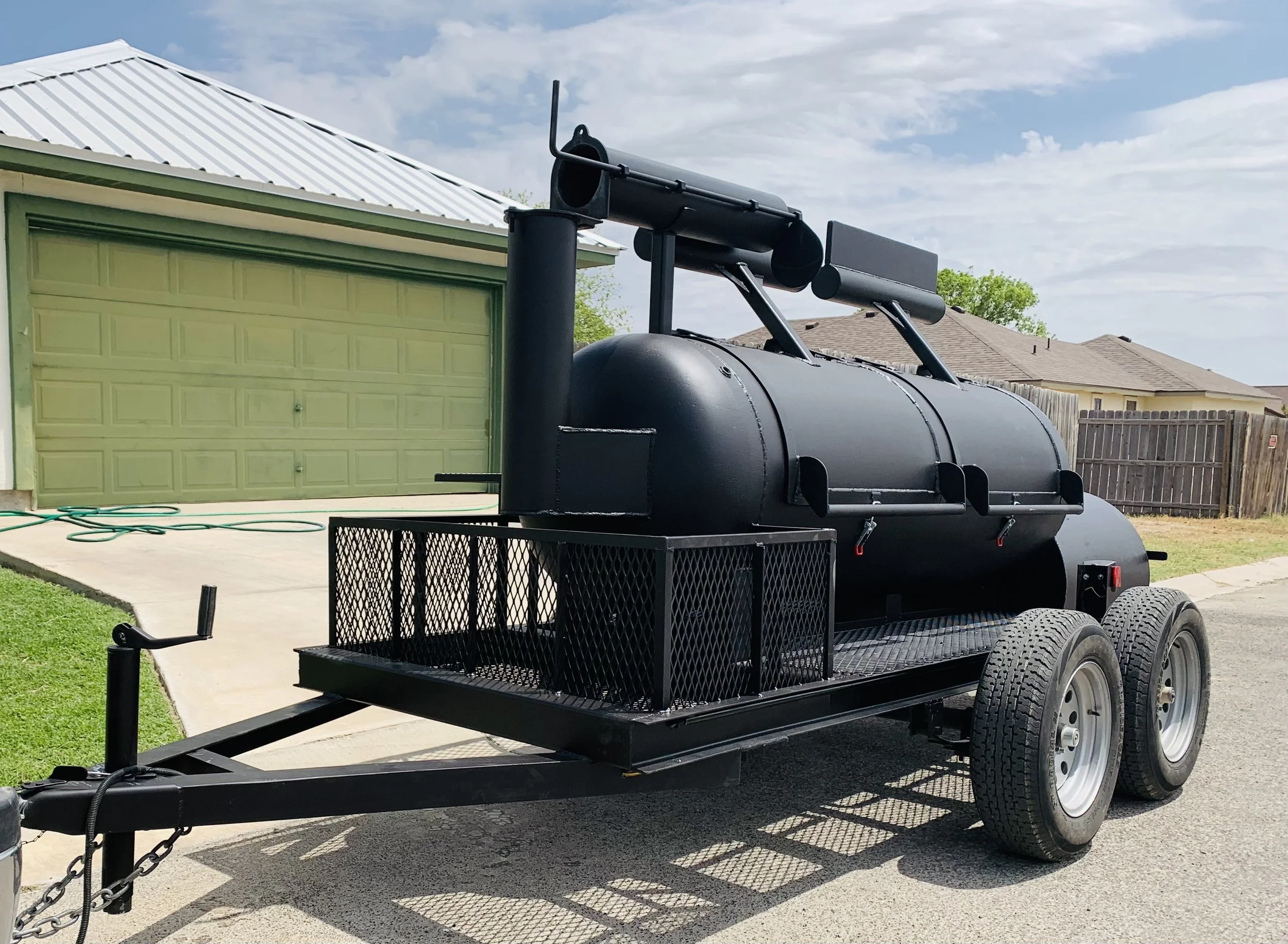 A black metal smoker grill on a trailer parked outside a house with a green garage door and a beige roof. The grill has a cylindrical body with a chimney on top and is attached to a trailer with two wheels and a hitch, with a black metal mesh shelf u