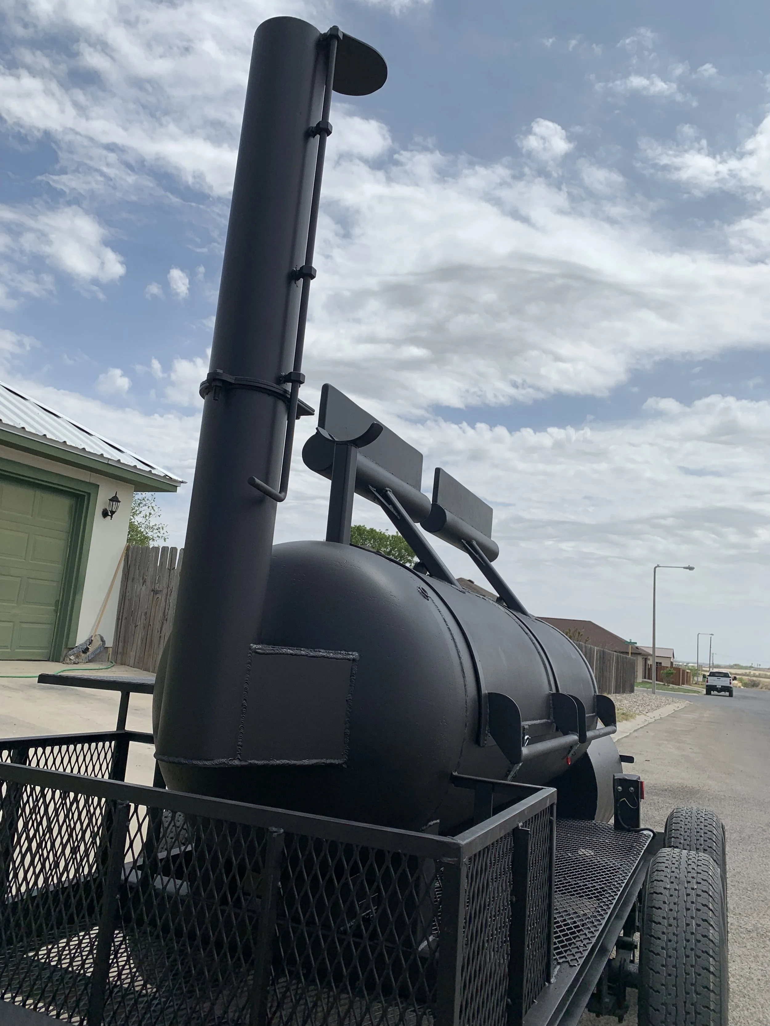 Black mobile smoker grill on a trailer with a large vertical chimney pipe, parked in a driveway near a house with a green garage door, under a partly cloudy sky.