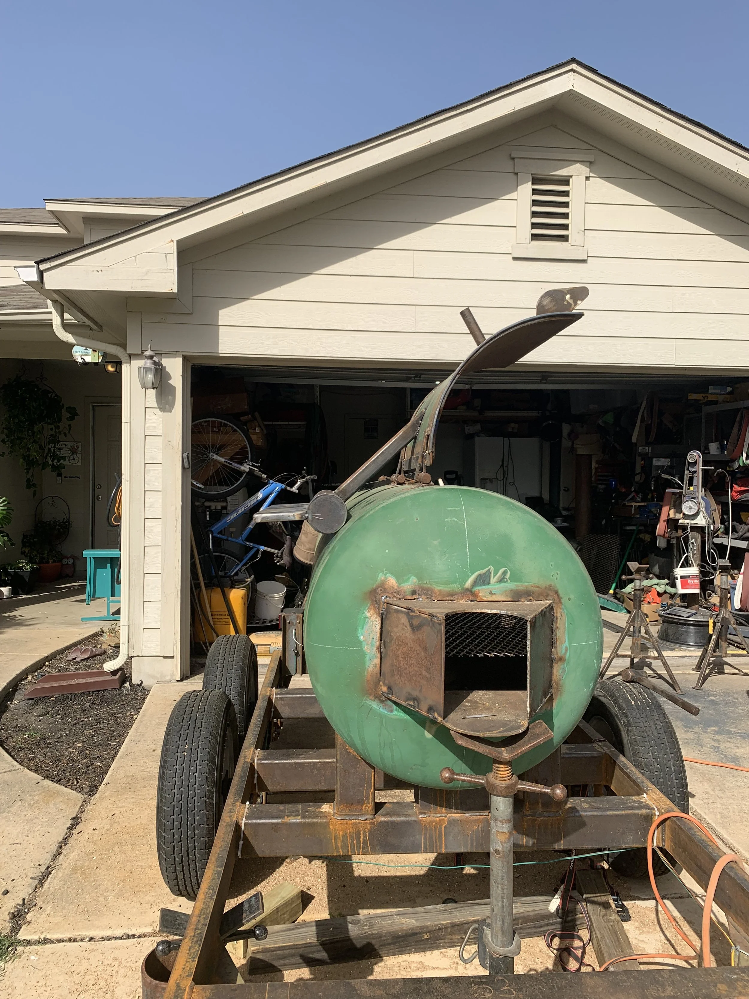 A vintage, rusty green tank on a flatbed trailer in a driveway, with a house in the background, various tools, and bicycle in the garage.