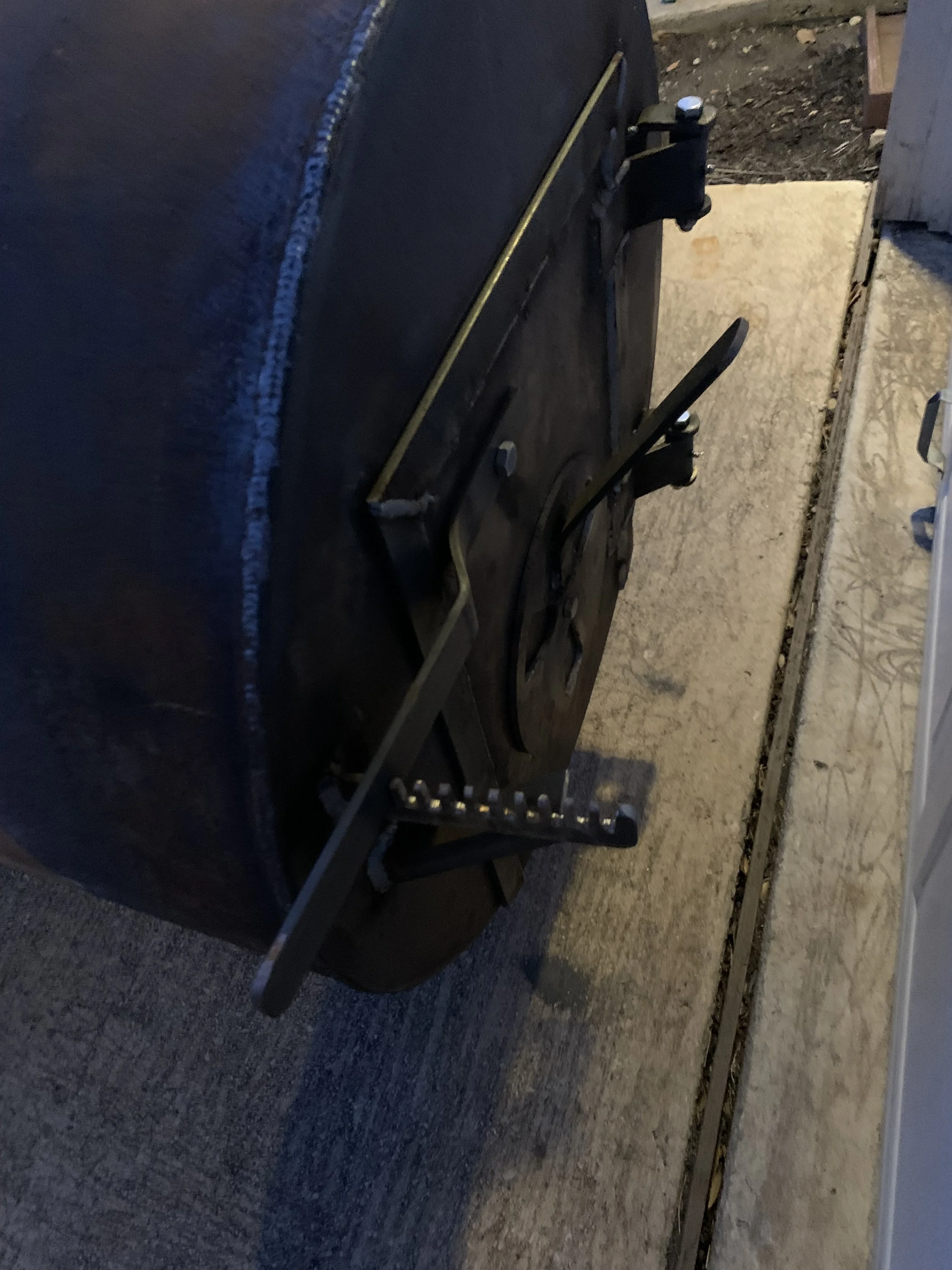 A black barbecue grill with a lid and side table, located outdoors on a concrete surface with gravel and dirt nearby.