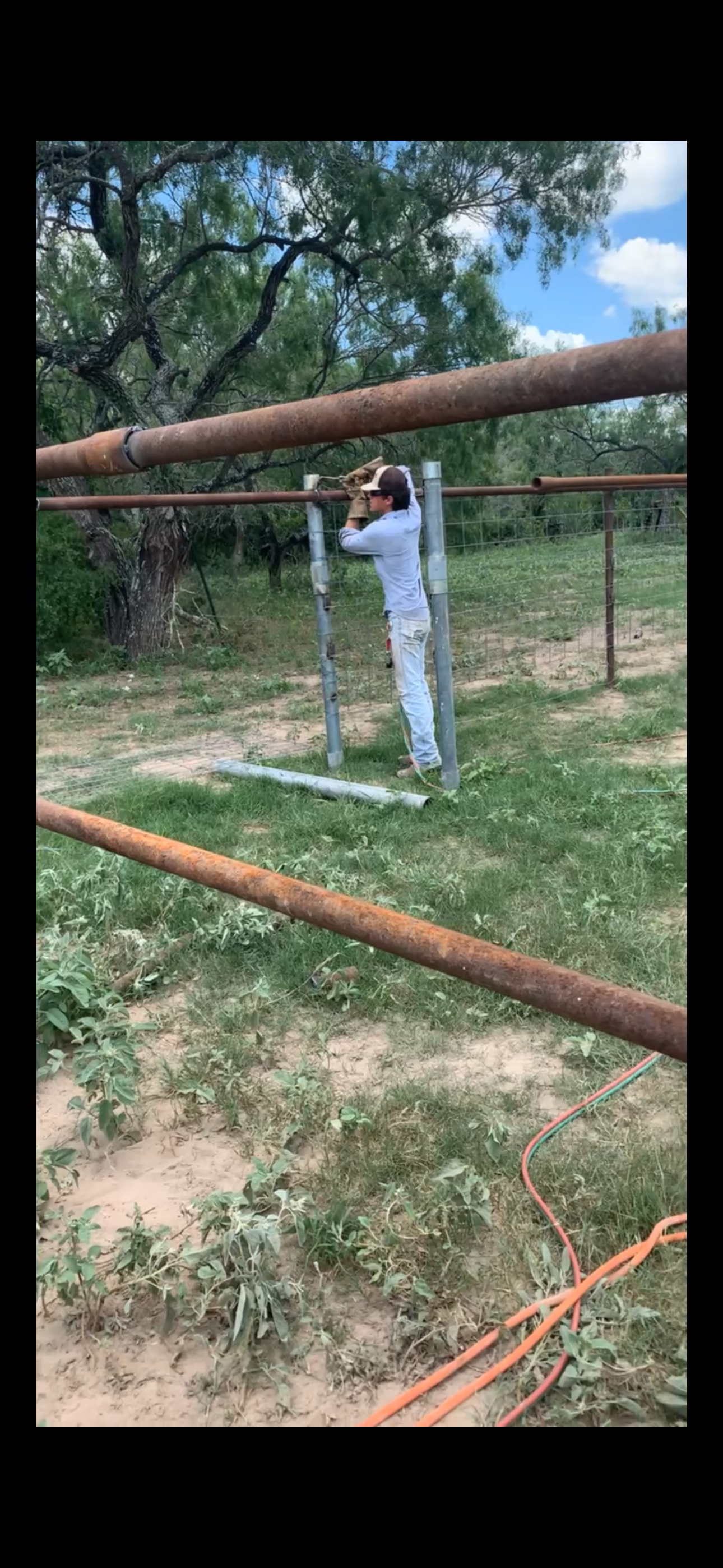A person wearing sunglasses, gloves, and a light gray long-sleeve shirt is working outdoors with a metal pipe, surrounded by trees, grass, and construction materials on the ground.