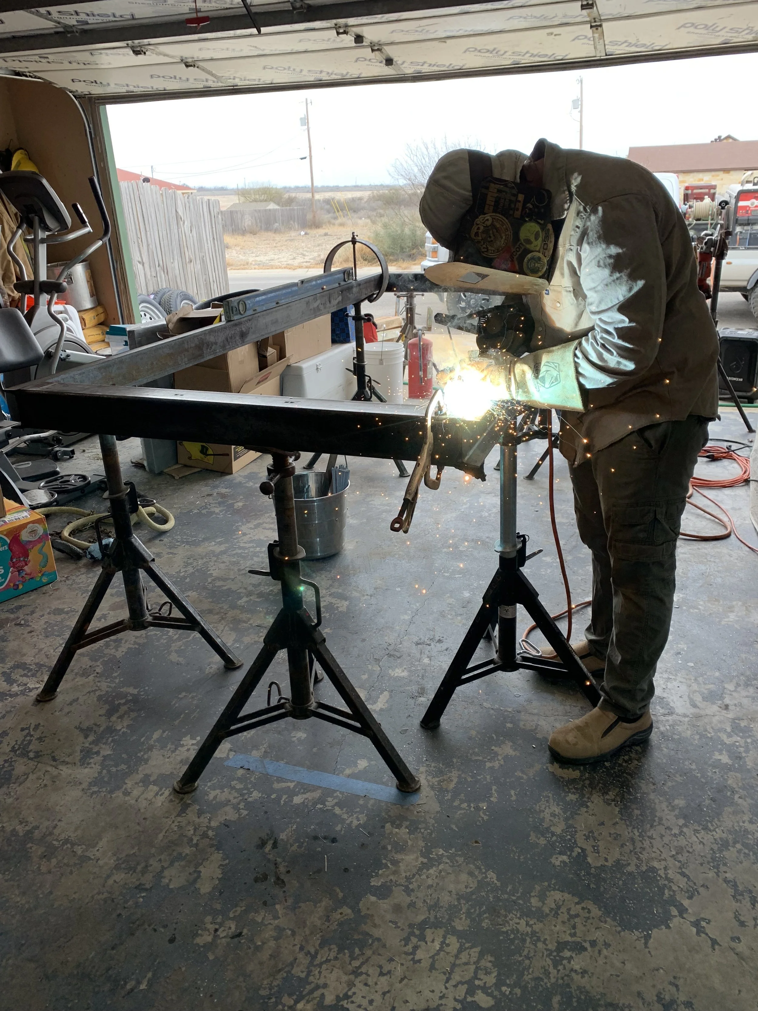 A person welding inside a garage with an open door, sparks flying from the welding process, various tools and equipment around, and an outdoor landscape visible in the background.