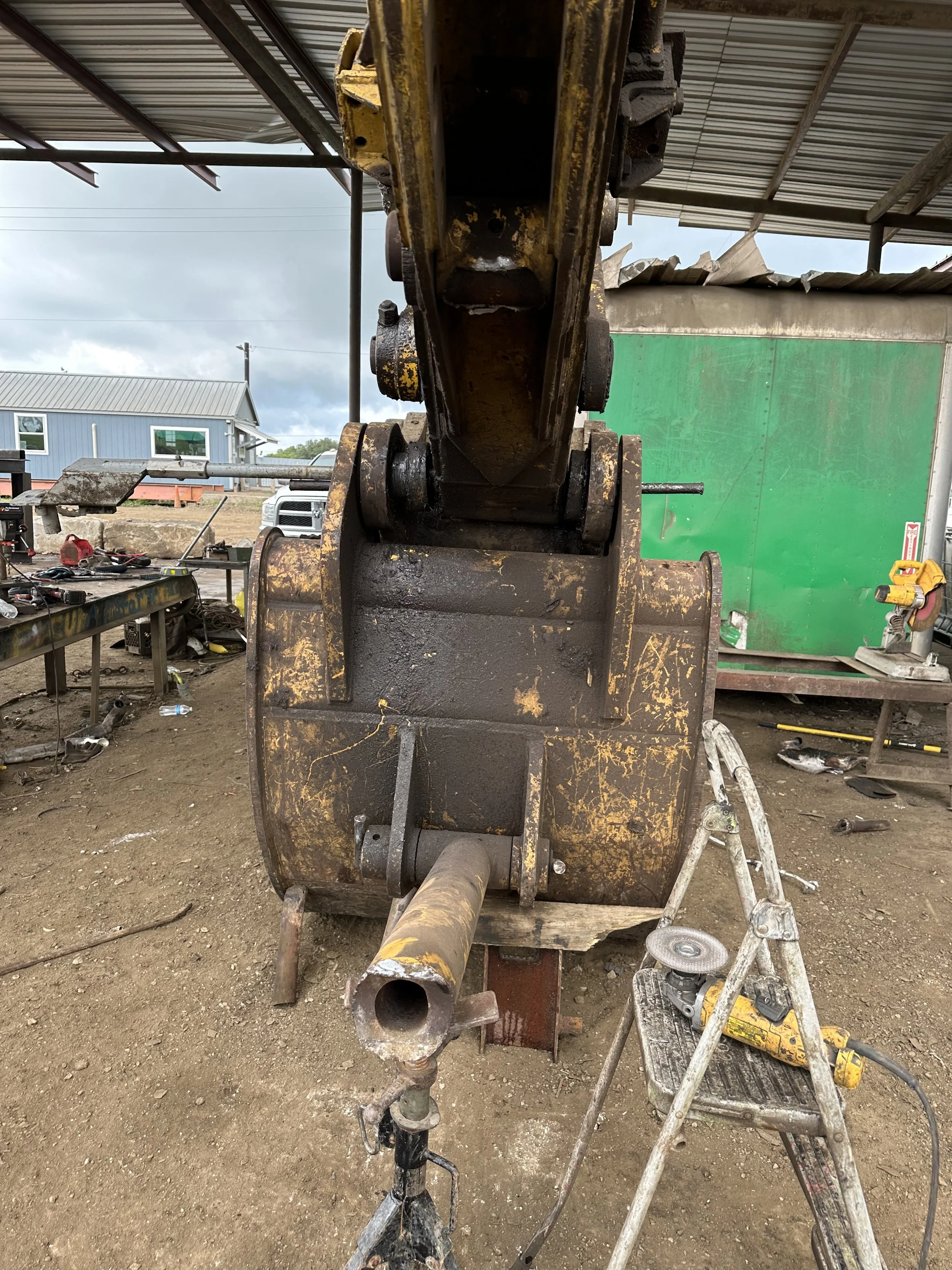 A construction site with a rusted excavator arm, tools, and equipment on a dirt ground under a metal roof, with a building and cloudy sky in the background.