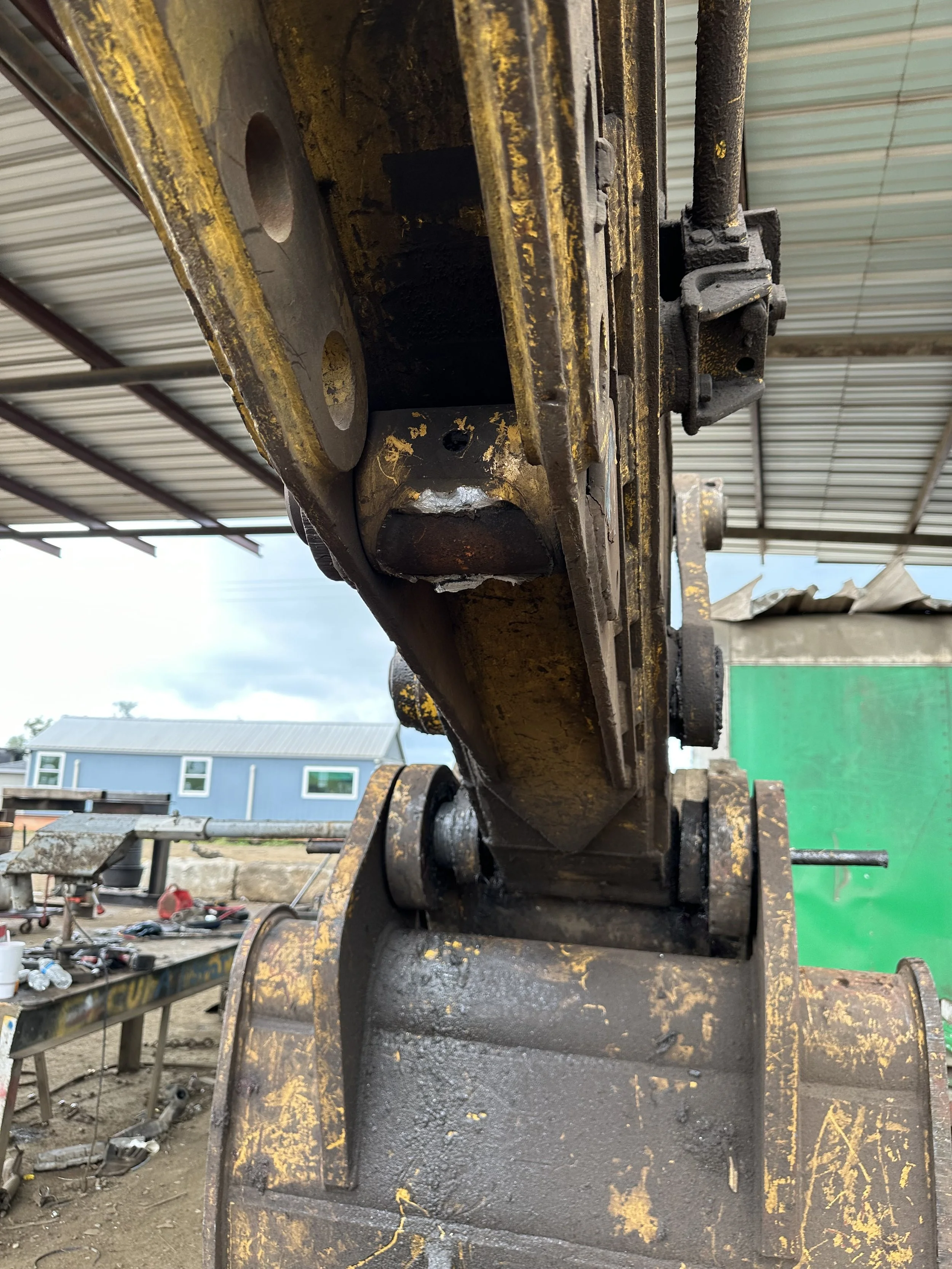 Close-up view of a rusty, yellow-painted construction excavator arm with visible wear and tear, image taken from beneath a metal roof in an outdoor construction site.
