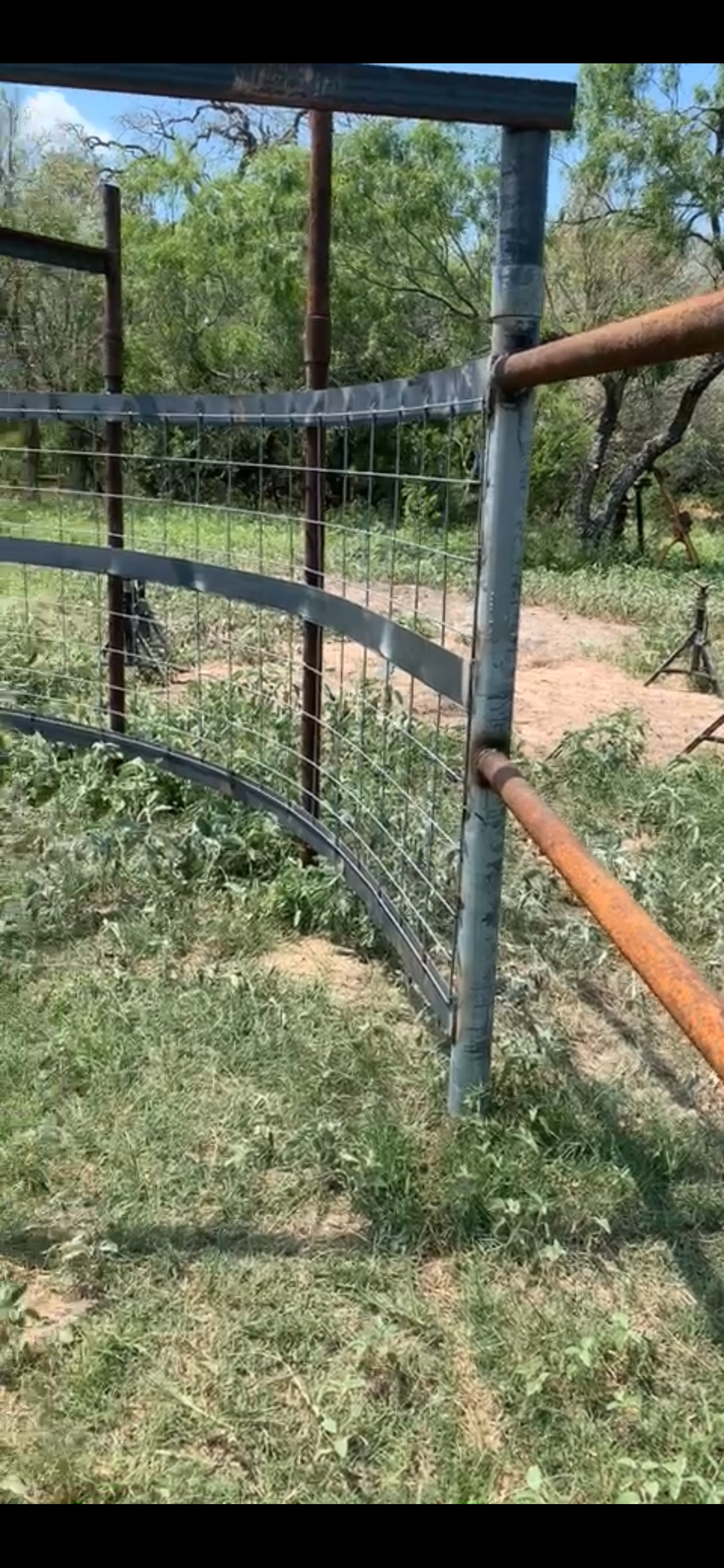 A metal fence with rusted horizontal bars and wire mesh panels, located in a grassy outdoor area with trees in the background.