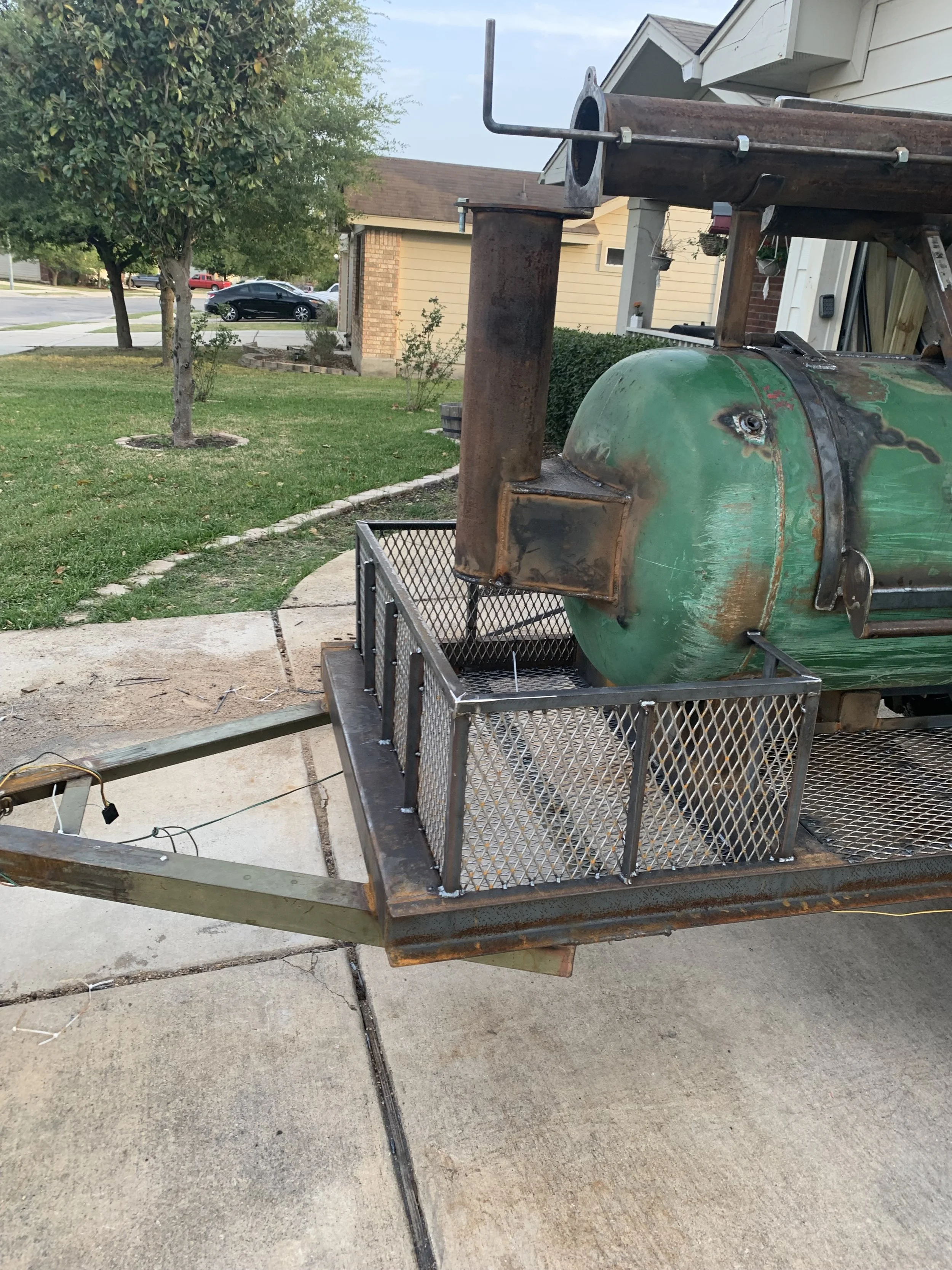 Part of a green grill with a rusty pipe attached, placed on a metal cart outdoors, with a tree, a house, and a street with cars in the background.