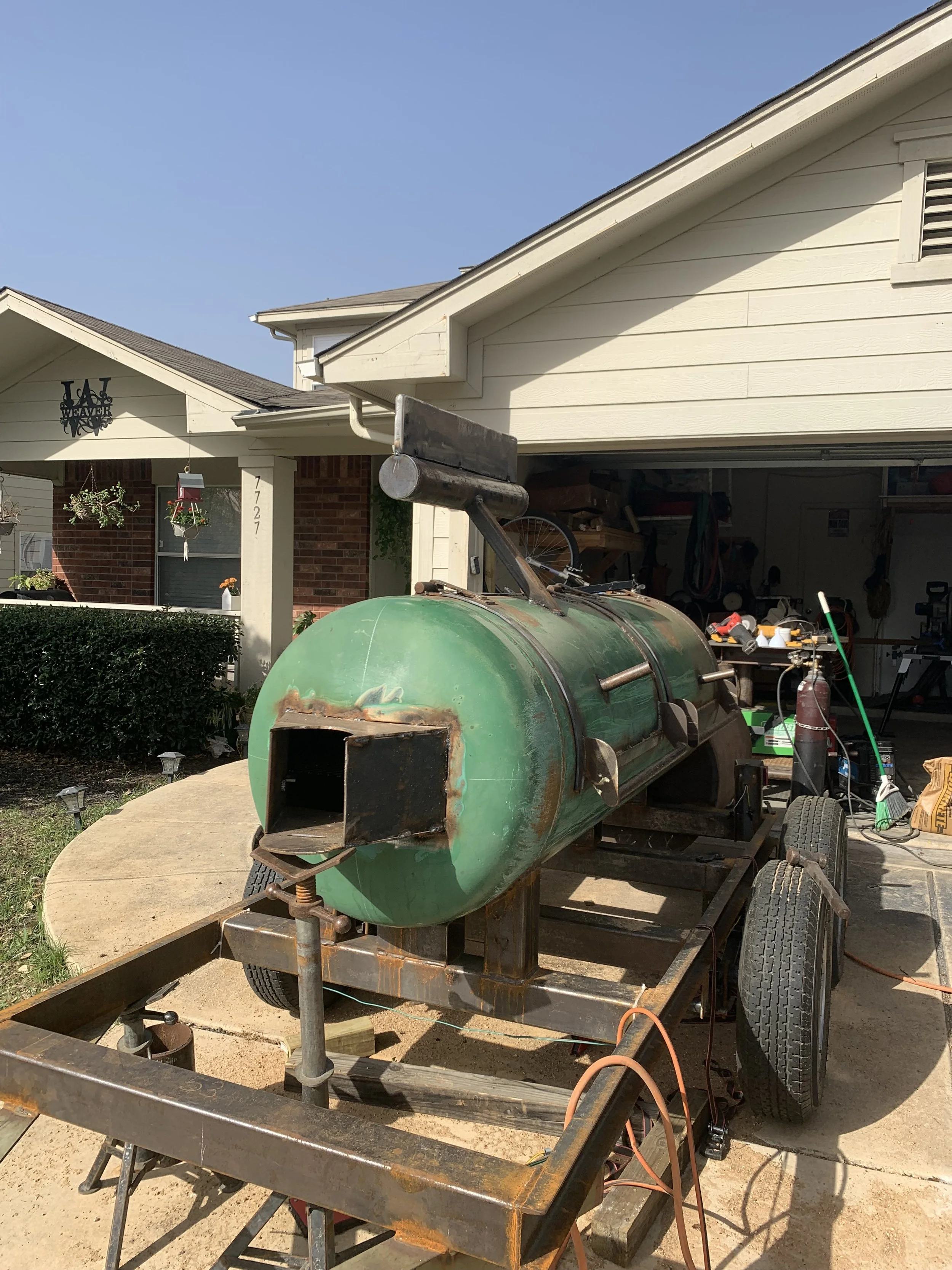 A rusty green metal boat on a trailer in a driveway in front of a house with a garage open, showing tools and equipment inside.