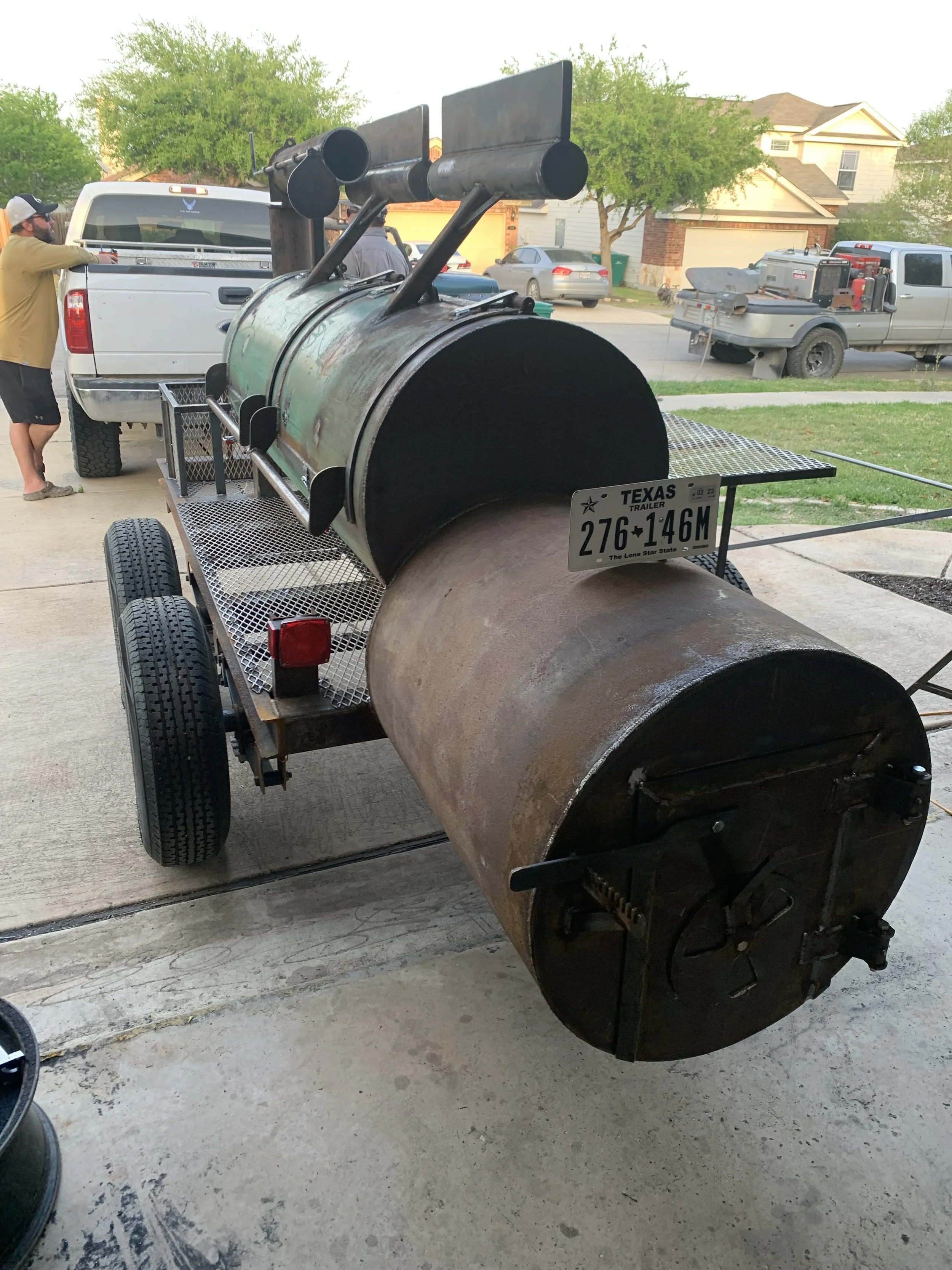 Large rusted metal smoker on a trailer, parked inside a garage with a concrete floor. Two people are near a white pickup truck in the background, with a neighborhood scene of houses, trees, and parked cars.