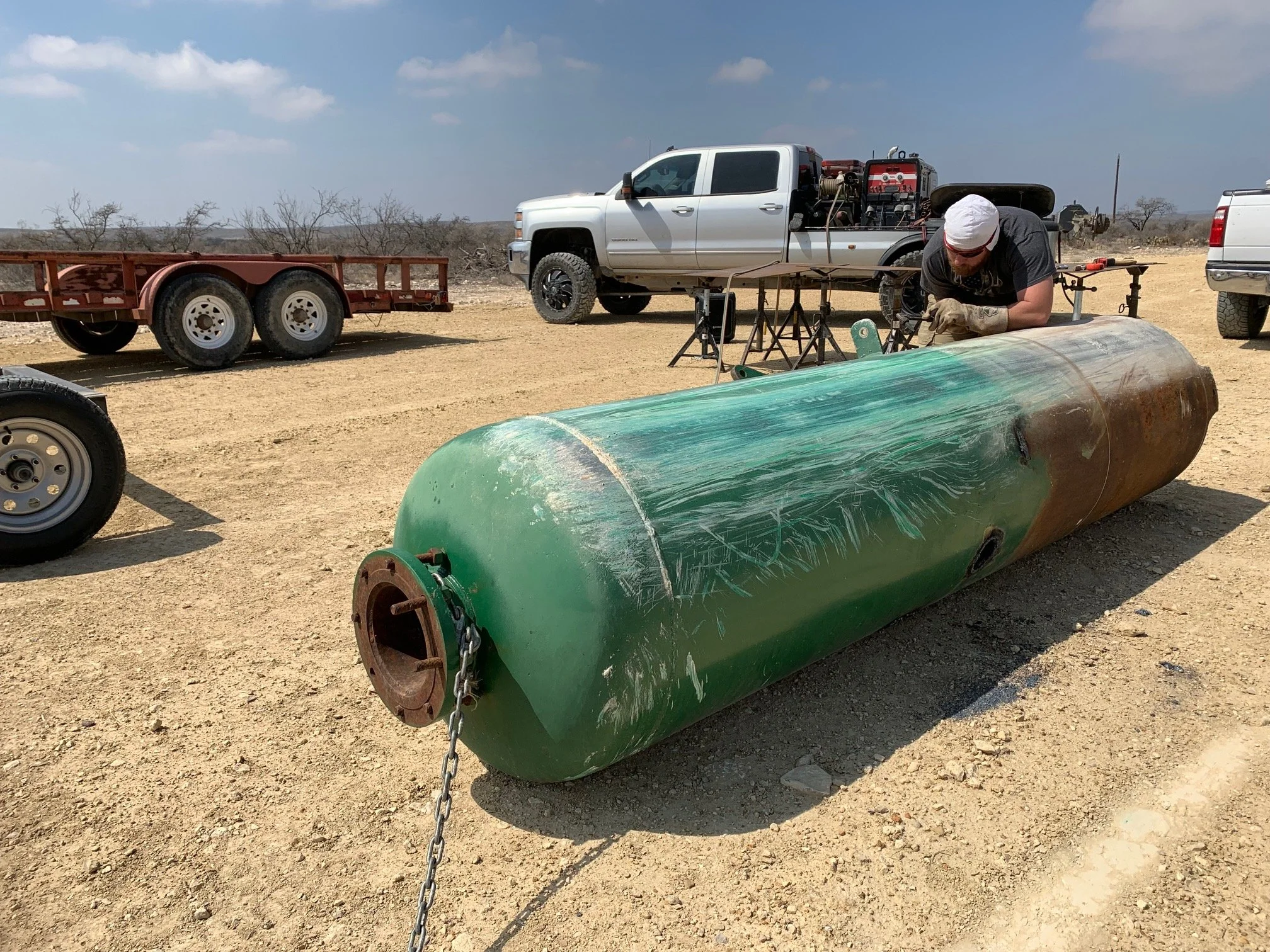 A man working on a green, rusted submarine torpedo with a chain attached to it, in a dry, dusty outdoor area with trucks and sparse, leafless trees in the background.