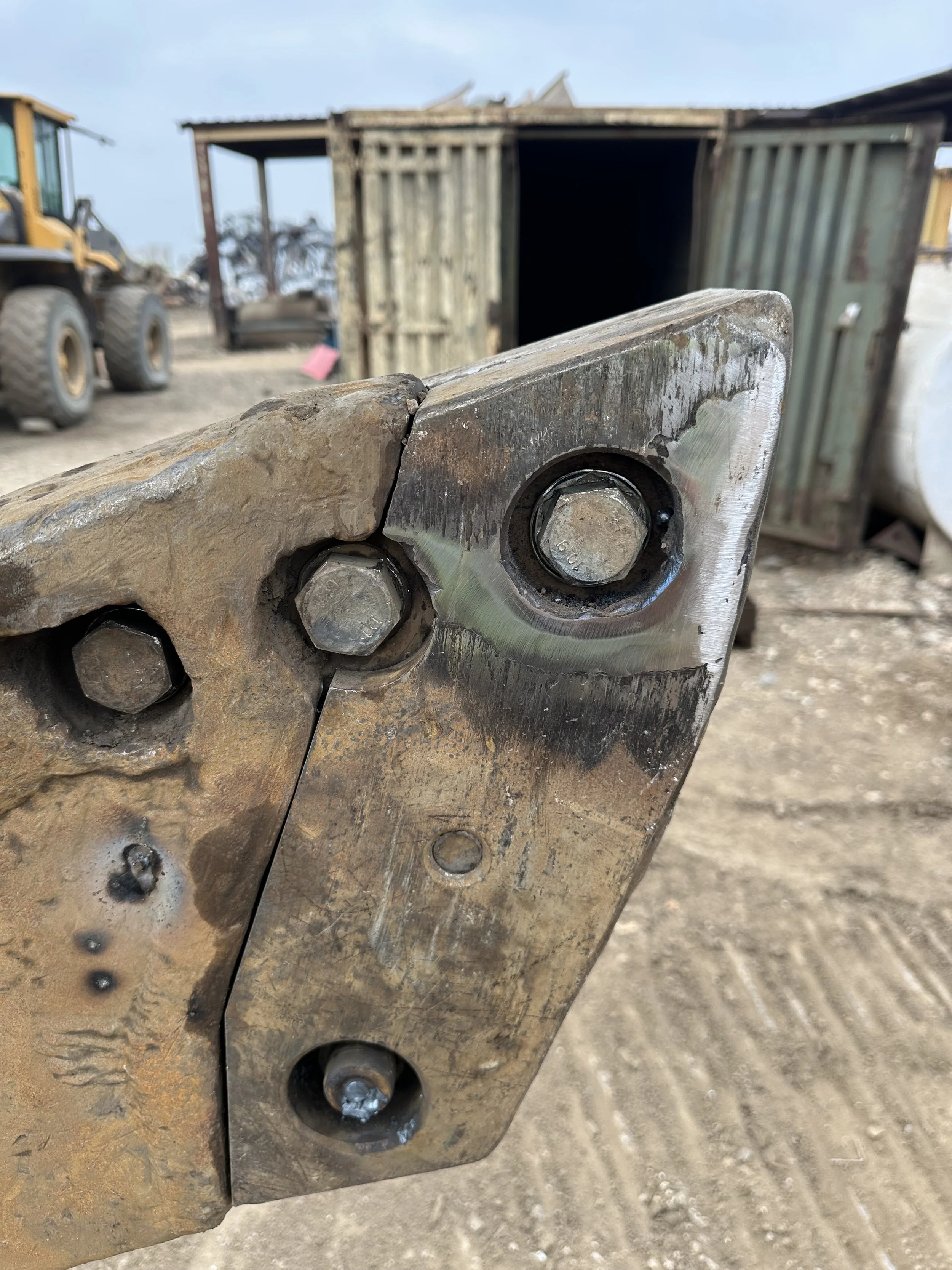 Close-up of a metal hinge or joint with large bolts, with a rusty and worn surface, part of a larger construction or mechanical assembly, outdoors with construction equipment and a wooden structure in the background.