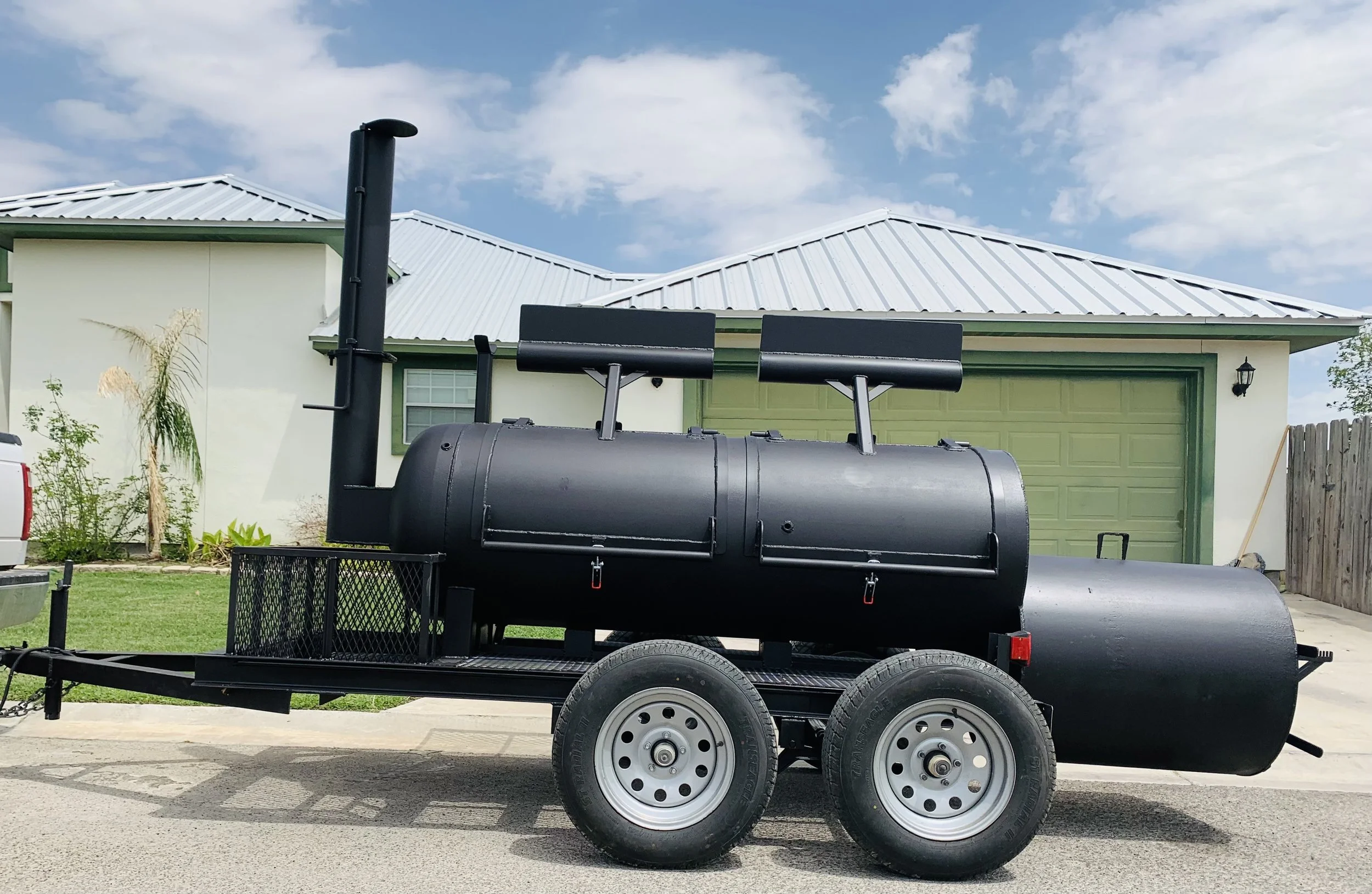 A black barbecue smoker on a trailer, parked in front of a house with a green garage door and a pale yellow wall. The house has a white metal roof and a black lantern outside. The yard has grass, plants, and a wooden fence on the right. The sky is pa