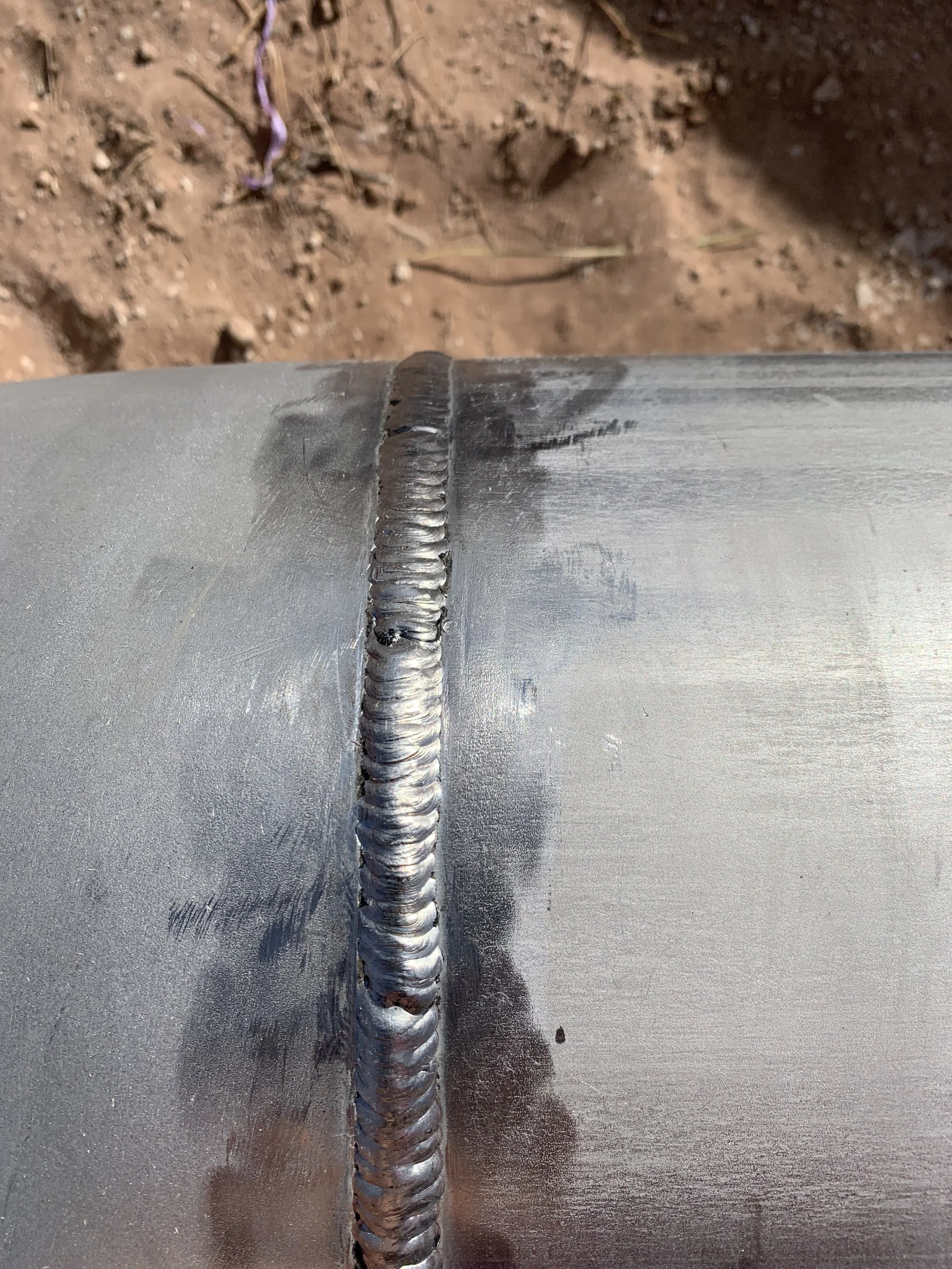 Close-up of a stainless-steel welded metal seam on a cylindrical metallic object, with a background of bare dirt ground. | John 6:9 Welding Company LLC | Sedalia, MO