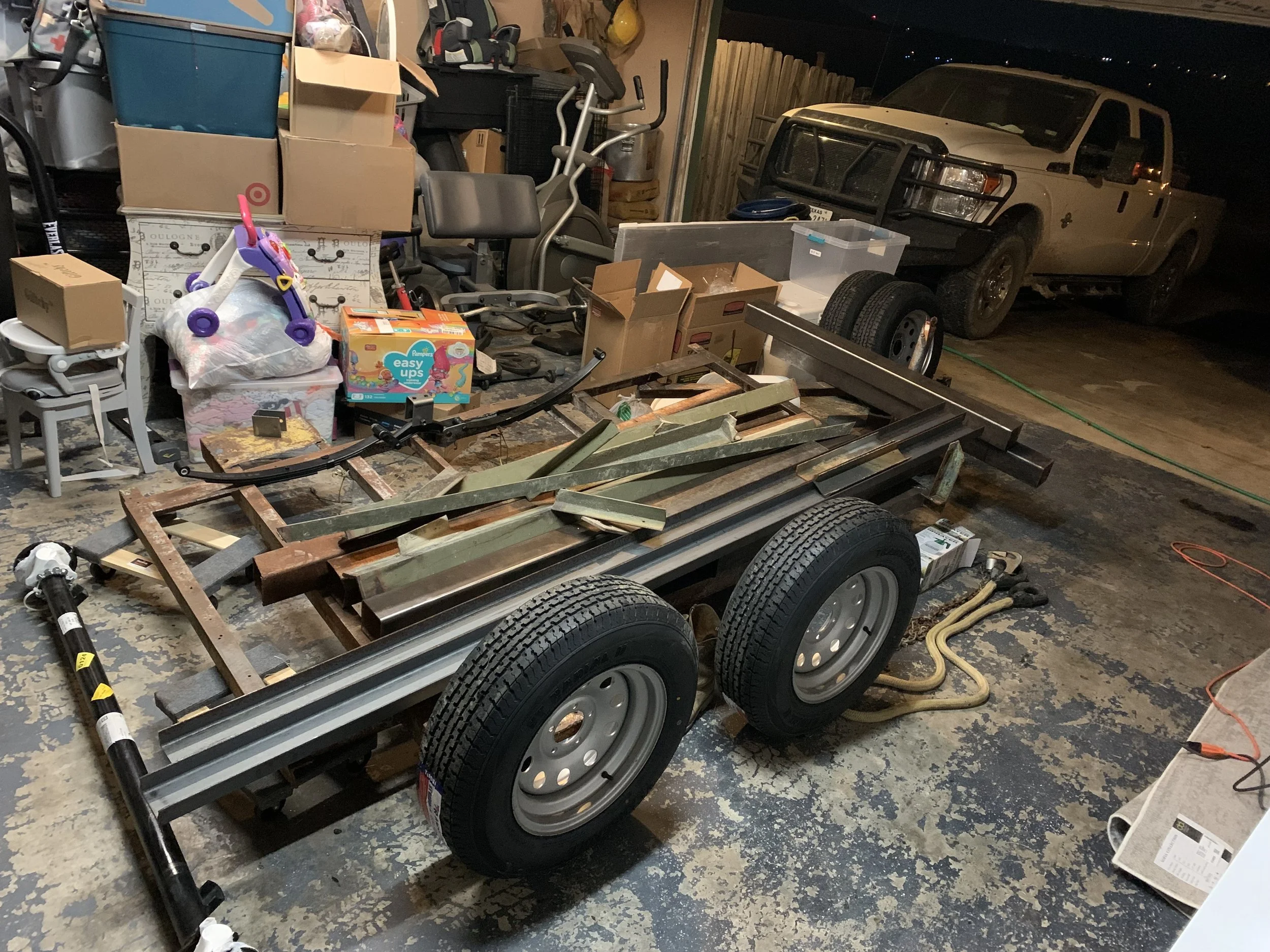 Garage filled with various items, including boxes, a children's tricycle, exercise equipment, and a large white van with a protective grille on the front. Items are scattered and there are tires and metal parts on the floor.