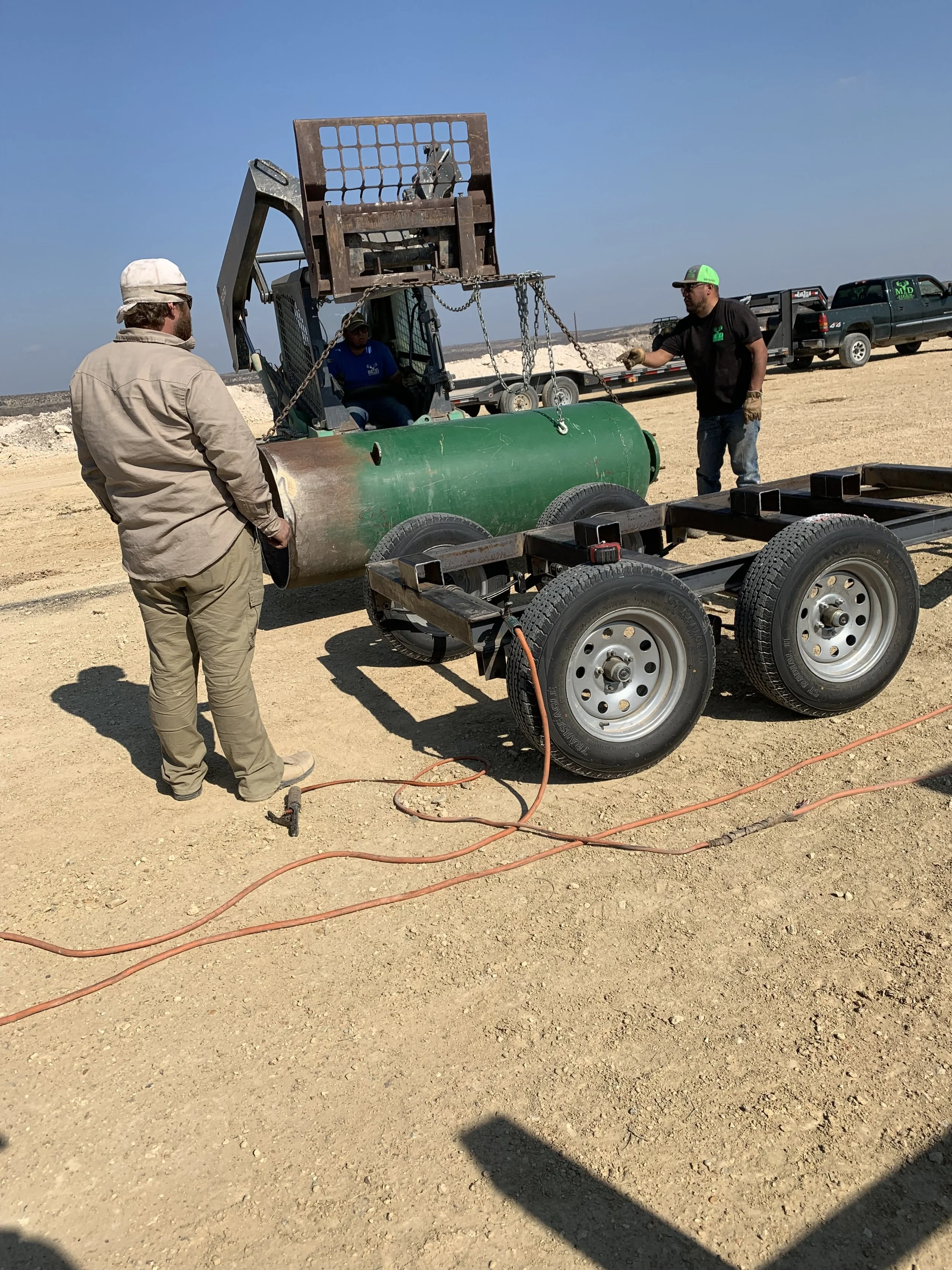 Three men working with a piece of industrial equipment on a dirt construction site. One man is standing with hands in pockets, another operating machinery, and the third sitting inside the machine. There are utility vehicles and trailers in the backg