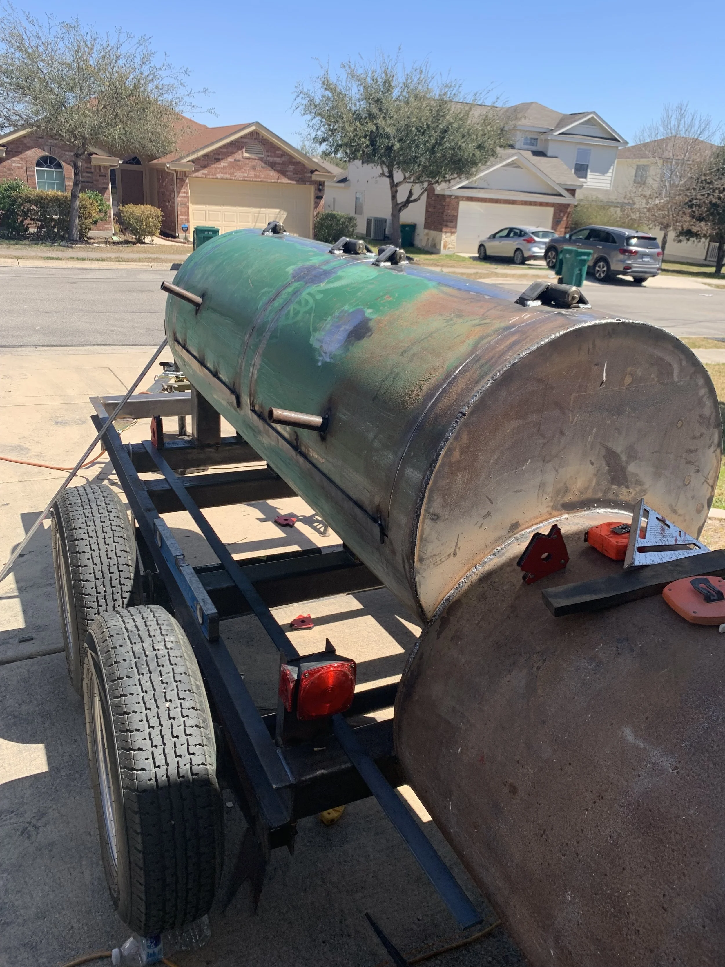 A large, cylindrical metal tank installed on a trailer, with a residential neighborhood in the background featuring brick and siding houses, trees, and parked cars.