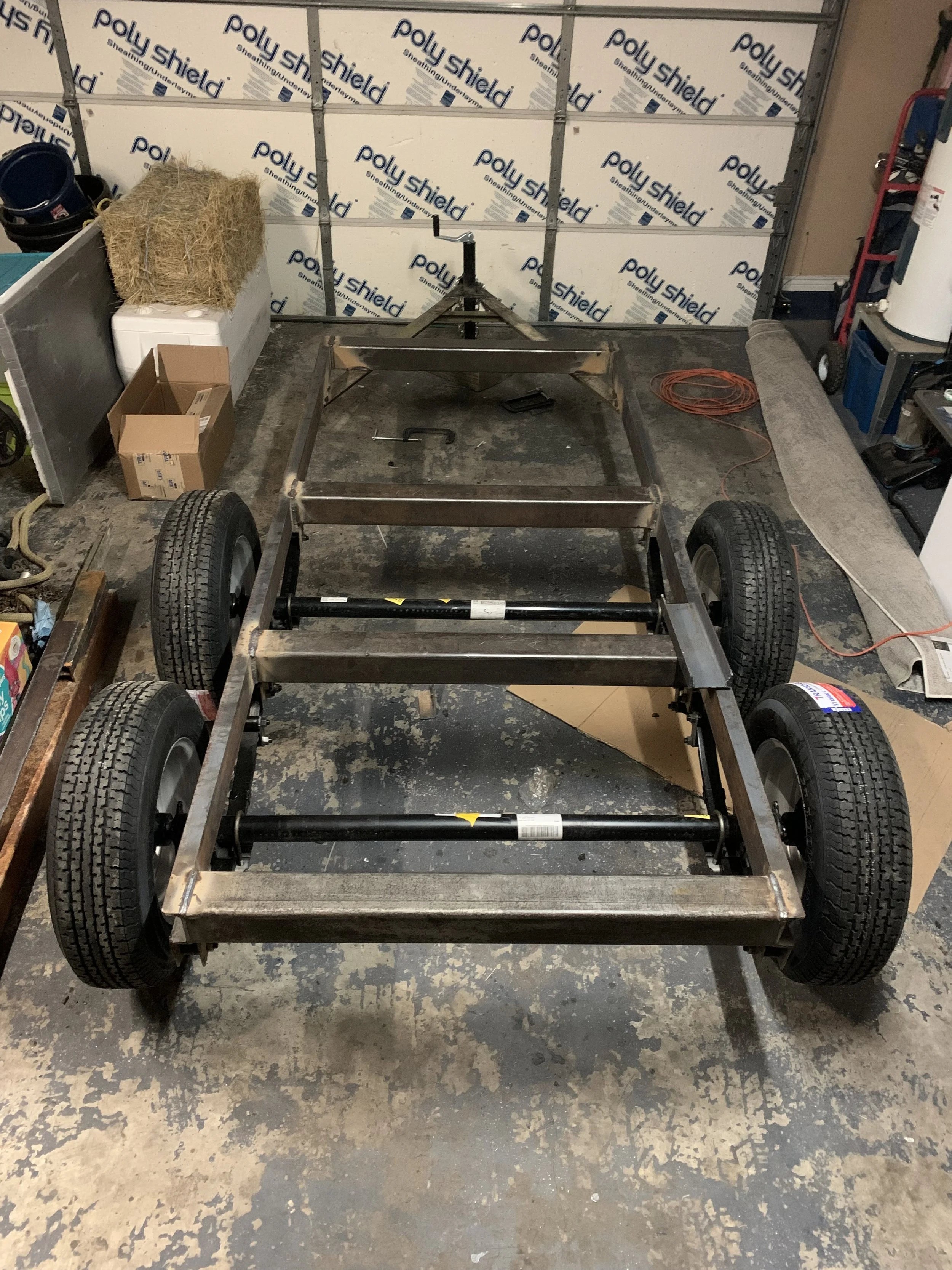 A metal frame structure on wheels, possibly a go-kart or small vehicle chassis, in a garage or workshop. The background features a wall with Polaris Shield insulation, and various tools, cables, and equipment are scattered around.