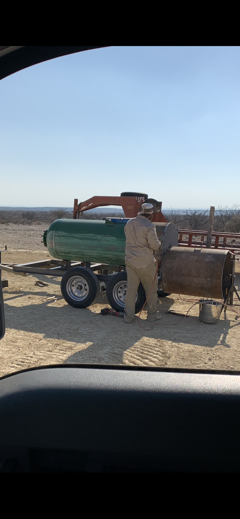 A person welding outdoors near a large green metal tank and rusty barrel, with a ladder and equipment on a dirt ground in a rural area under a clear blue sky.