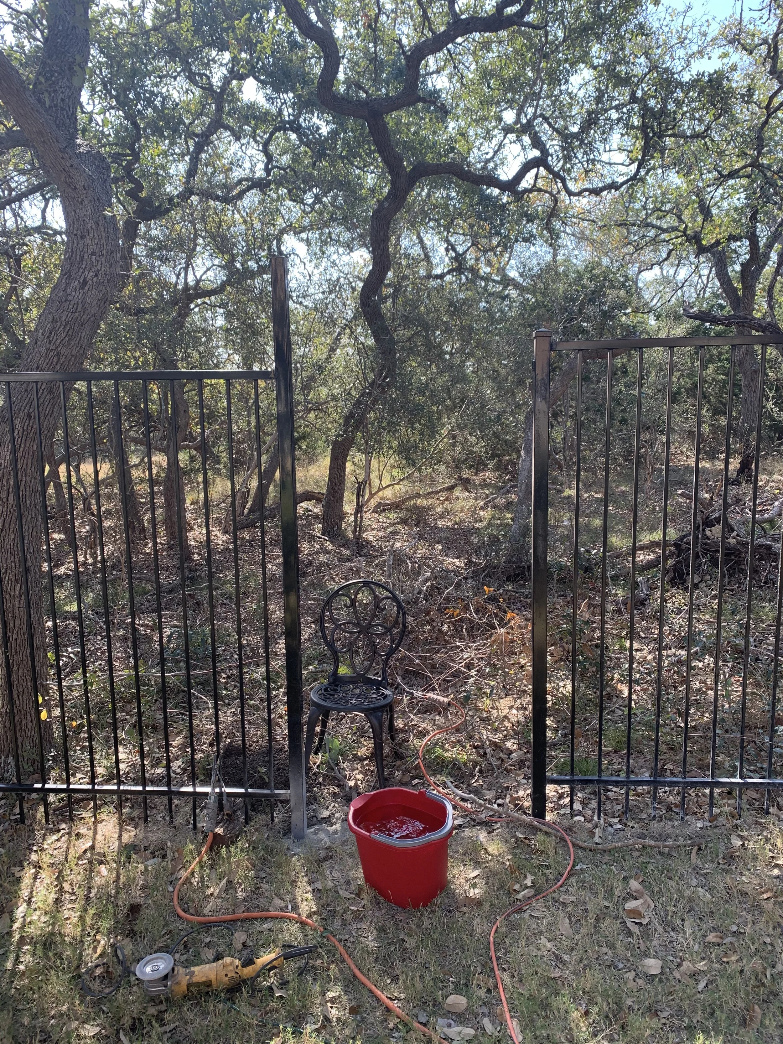 A metal garden gate opens to a backyard or natural wooded area with trees and dry grass. There is a black chair with decorative circular patterns on the backrest, and gardening tools including an orange extension cord, a small power tool, and a red b