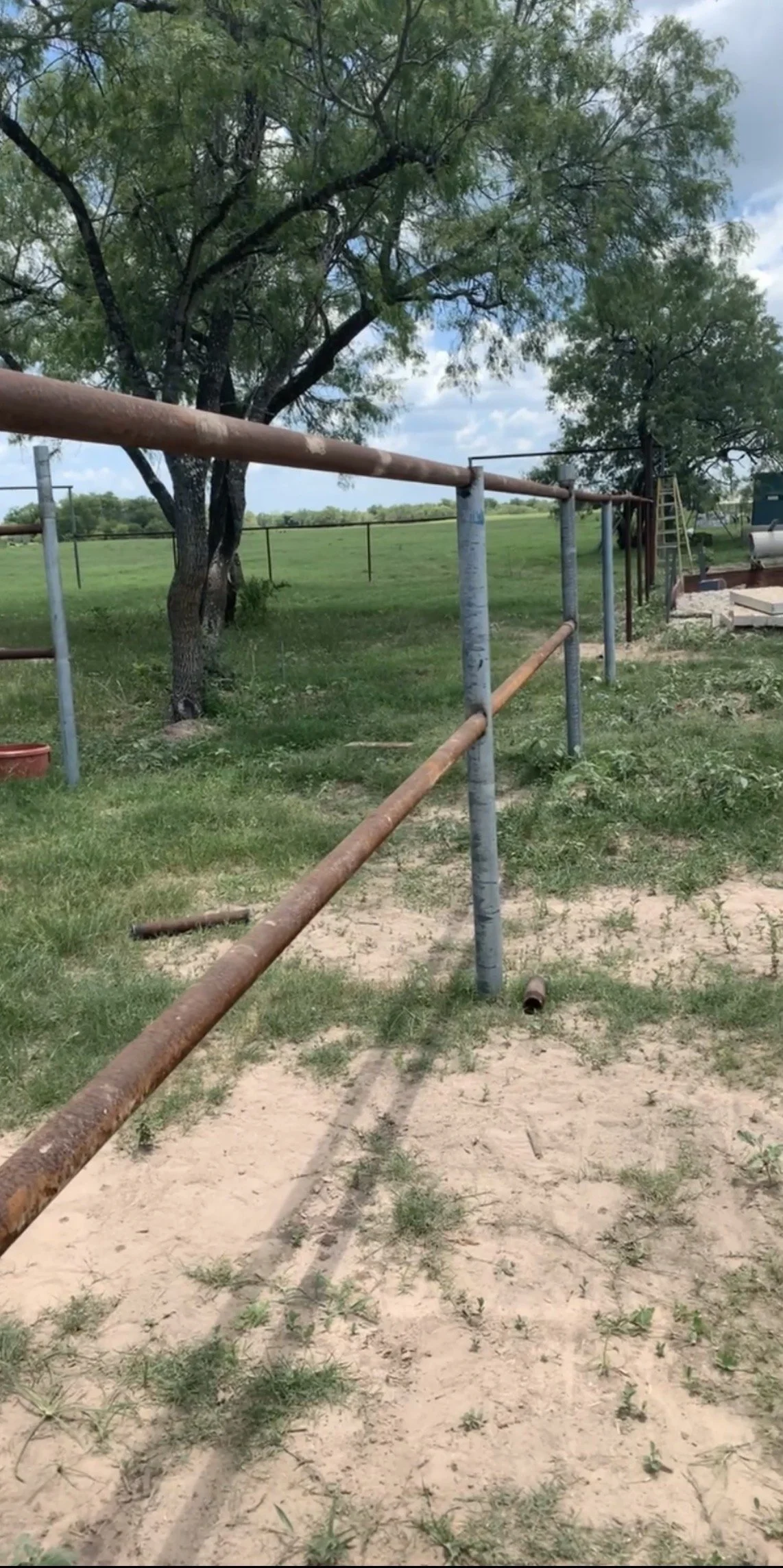 A dirt and grass area with rusty metal pipes and posts forming a makeshift fence or barrier, with large trees in the background and a cloudy sky.
