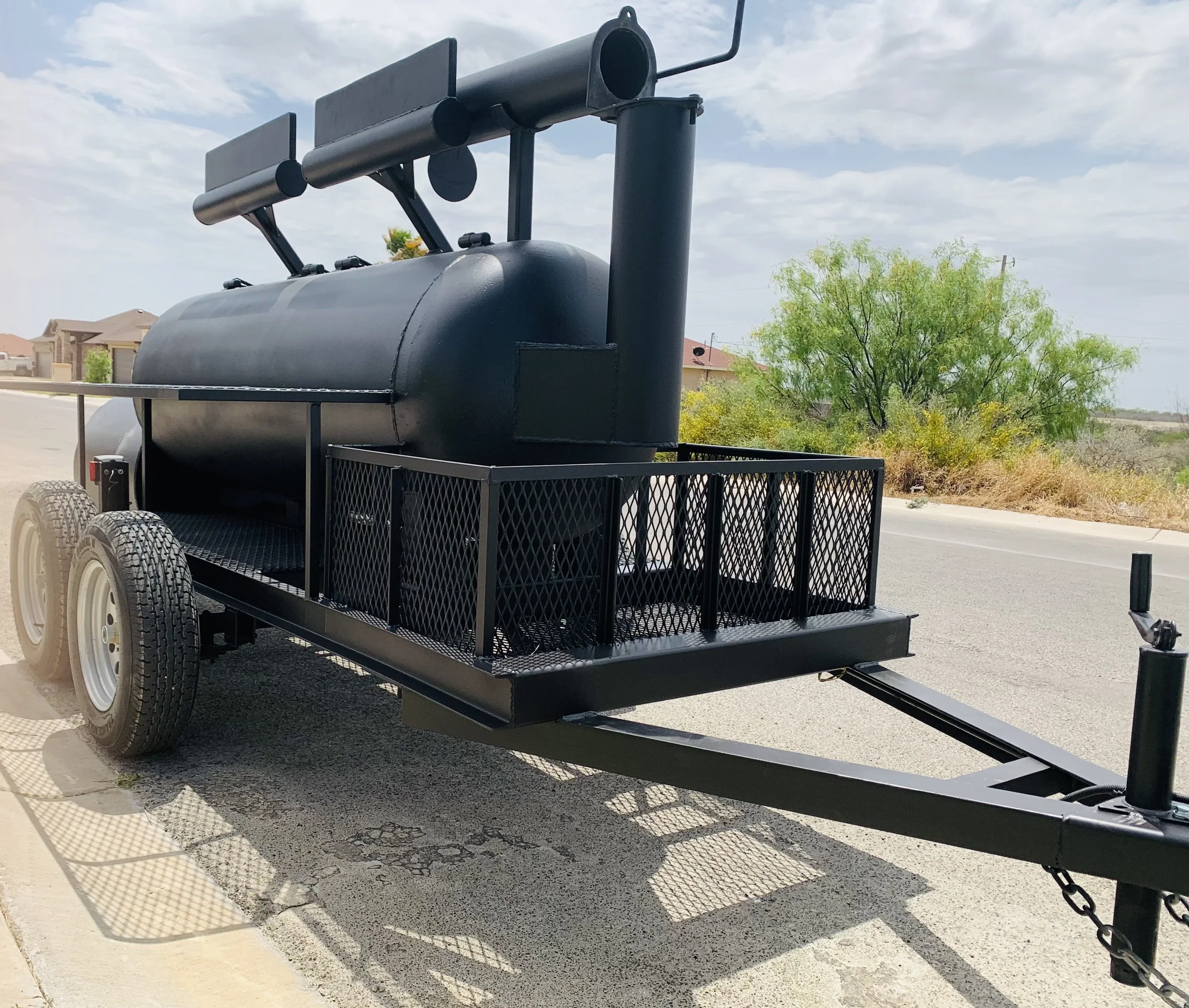 A large black smoker grill on a trailer with dual wheels parked on a paved surface, casting shadows on the ground, with trees and houses in the background and a partly cloudy sky.