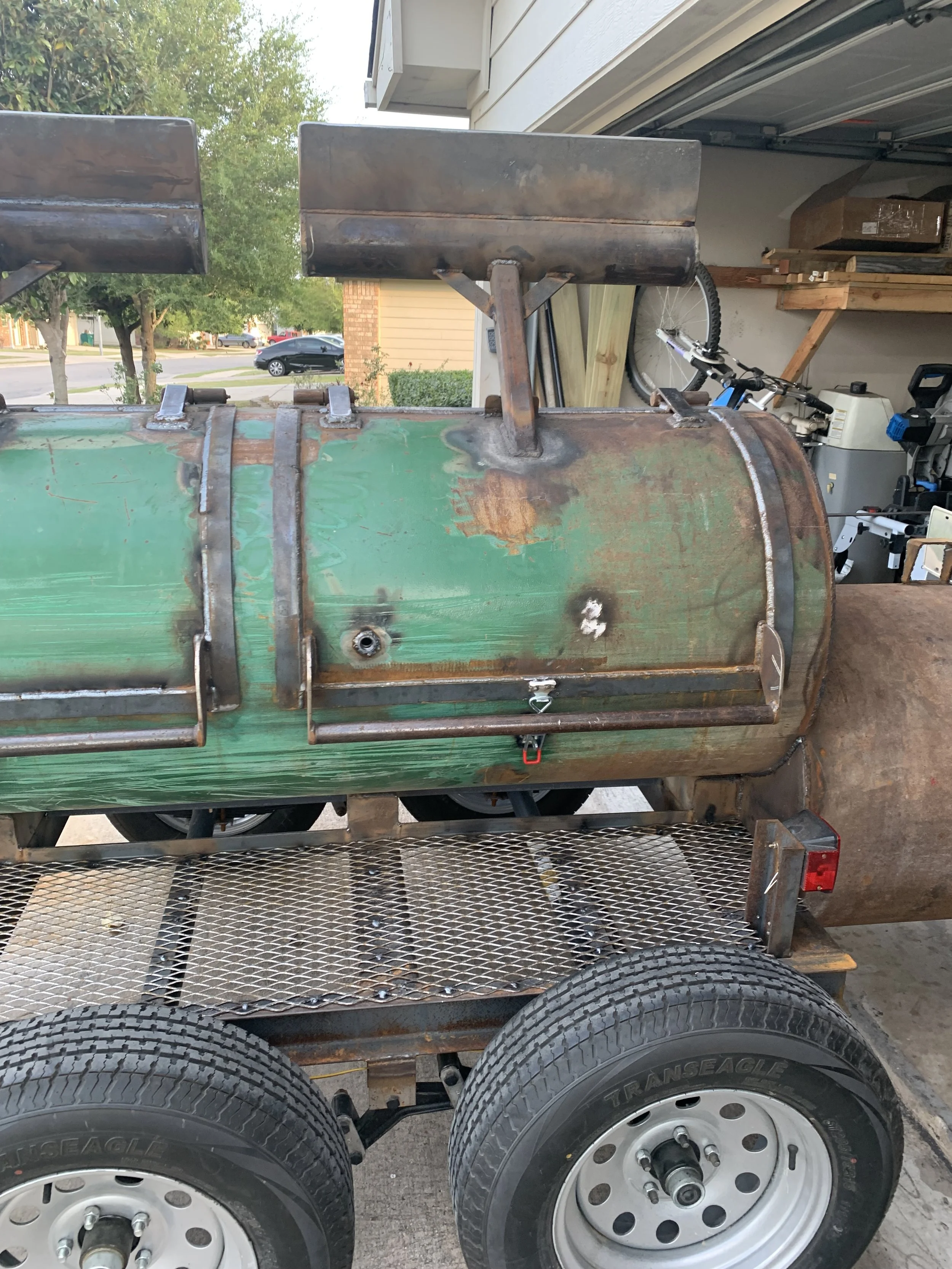 Close-up of a green metal barbecue smoker on a trailer, with parts of the trailer, tires, and a garage in the background.