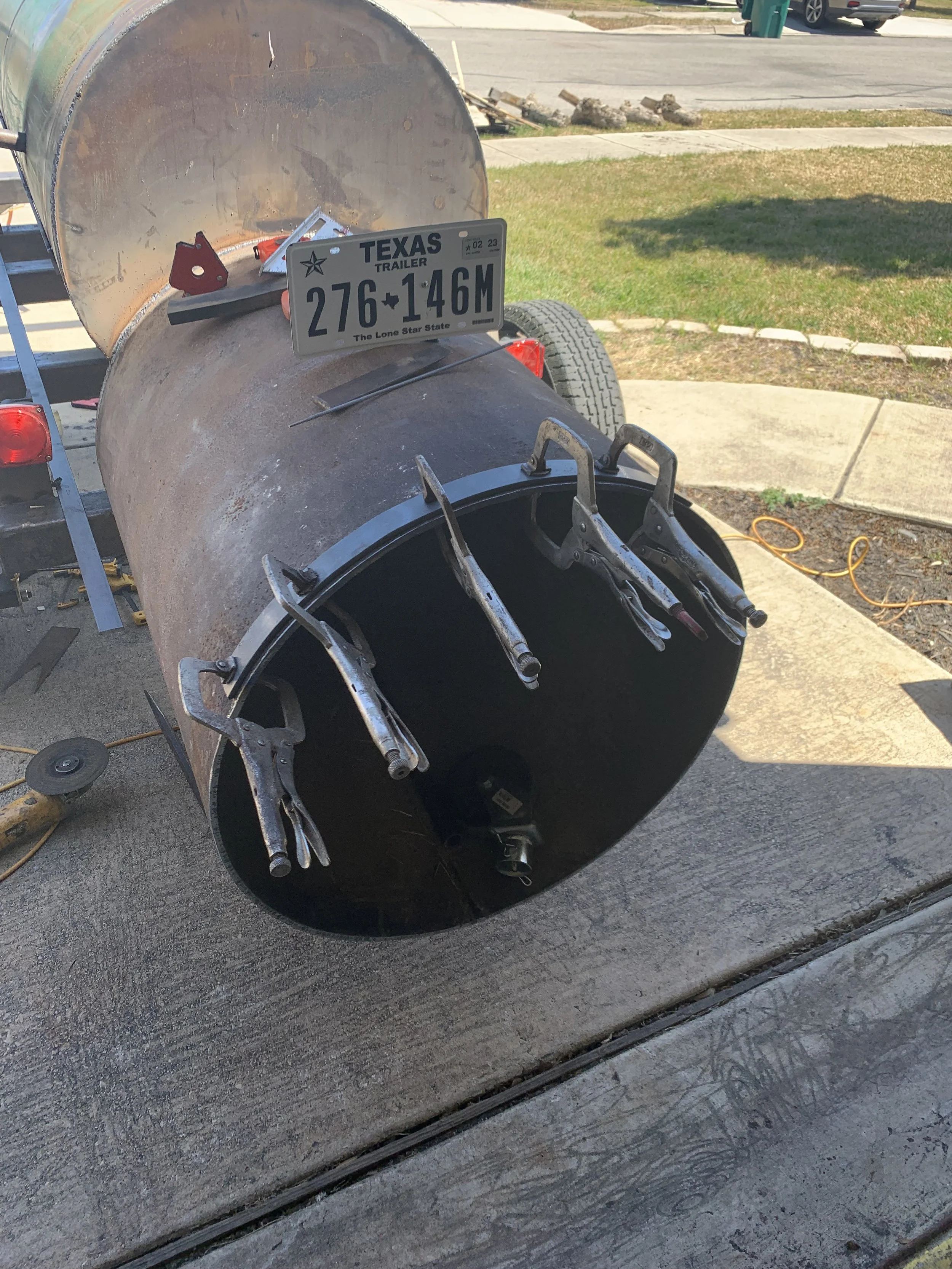 A large metal drum trailer with various clamps and tools attached, parked on a driveway near a sidewalk and a street with grass and a few cars in the background. A Texas license plate with the number 276-146M is attached to the trailer.