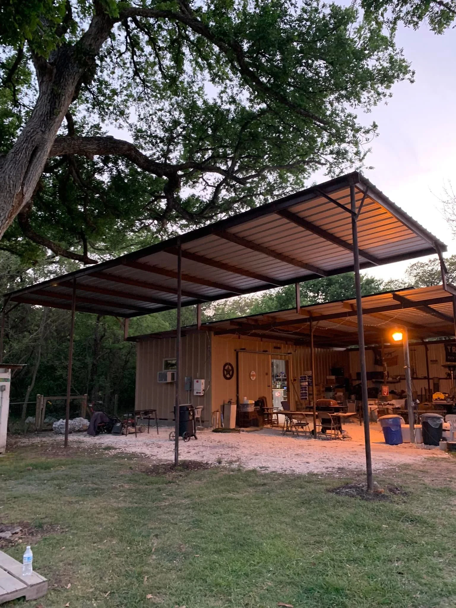 An outdoor scene showing a building with a metal roof and surrounding furniture, with trees and a cloudy sky in the background, under dim evening lighting.