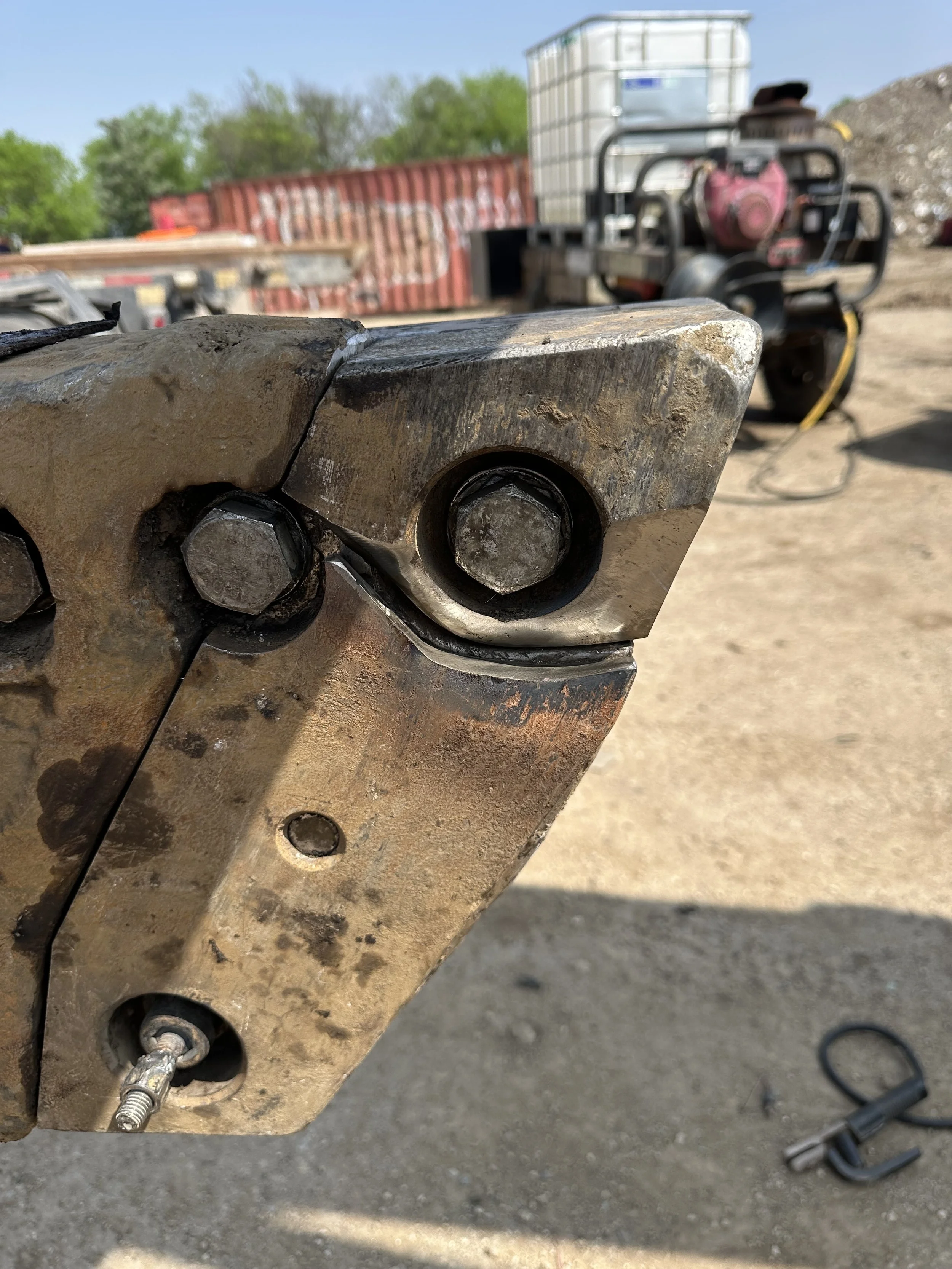 Close-up of a rusty metal industrial component with bolts and an attached hydraulic or pneumatic fitting. In the background, there are construction or industrial tools, a red and white container, and trees under a clear sky.