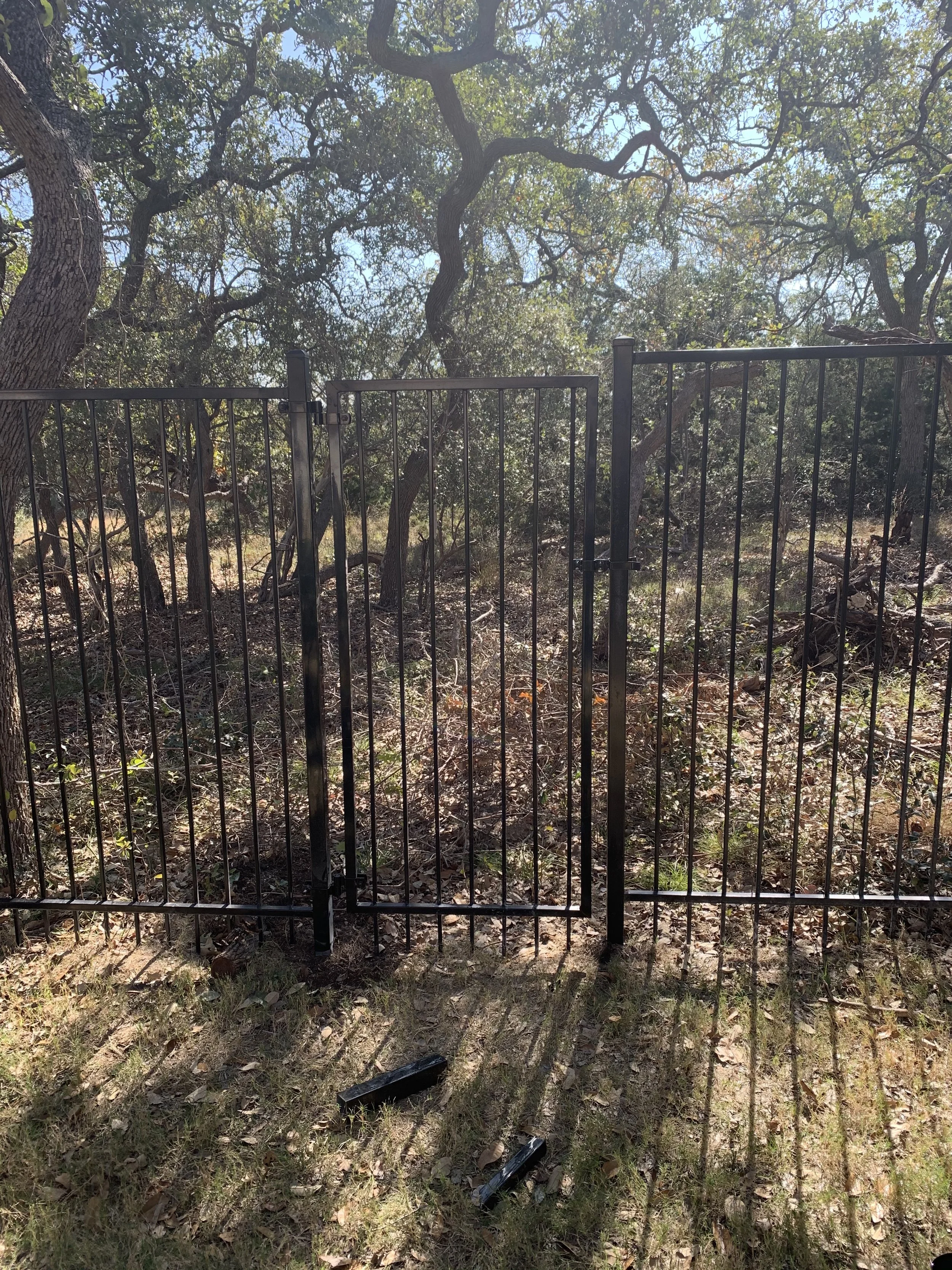 A black metal fence with a partially open gate is situated in front of a densely wooded area with tall trees, green leaves, and a partly cloudy sky visible through the branches.