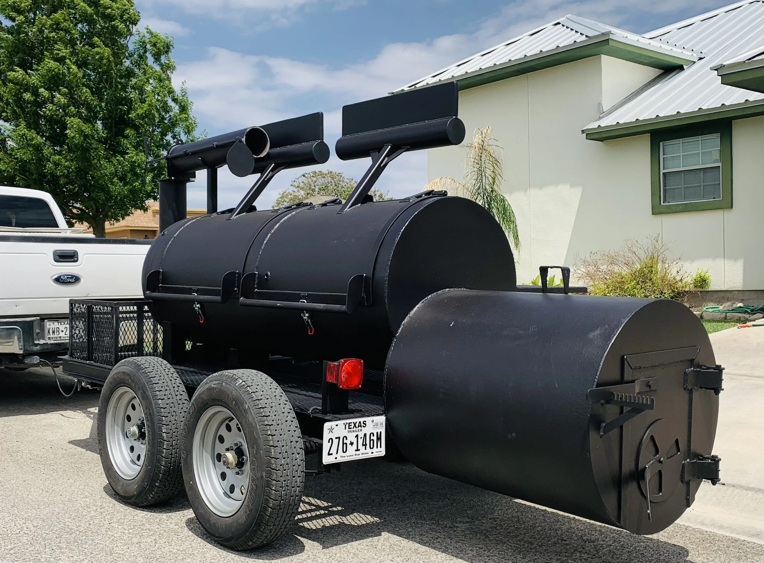 A black barbecue smoker on a trailer with a Texas license plate, parked on a driveway in front of a house with a white exterior and green trim. A white pickup truck is partially visible on the left side, and there is a large green tree and a cloudy s