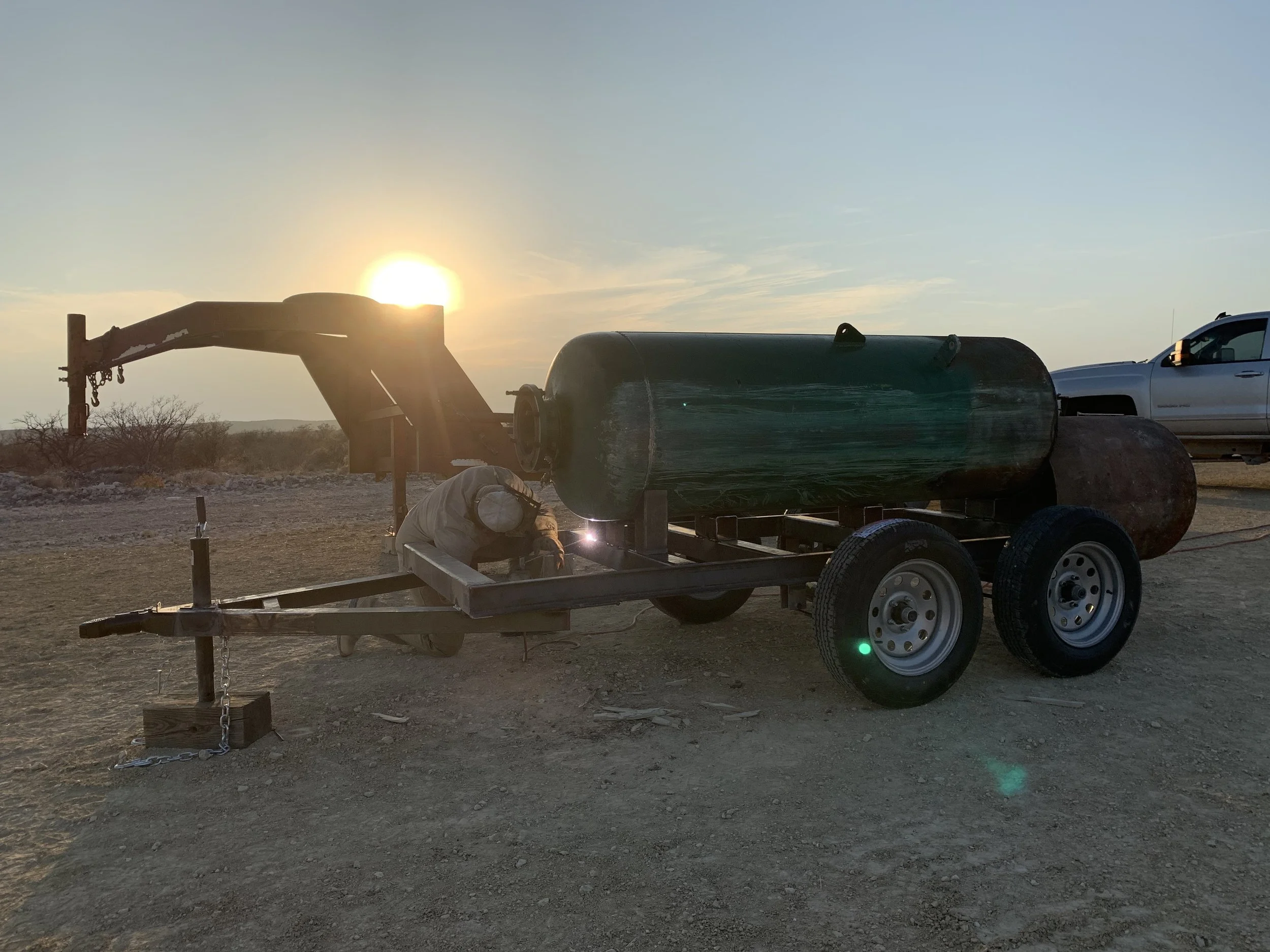 A flatbed trailer carrying a small green boat with a black cover, a gray pickup truck in the background, and a sunset in the sky. The trailer is parked on a dusty, open terrain with some sparse bushes.