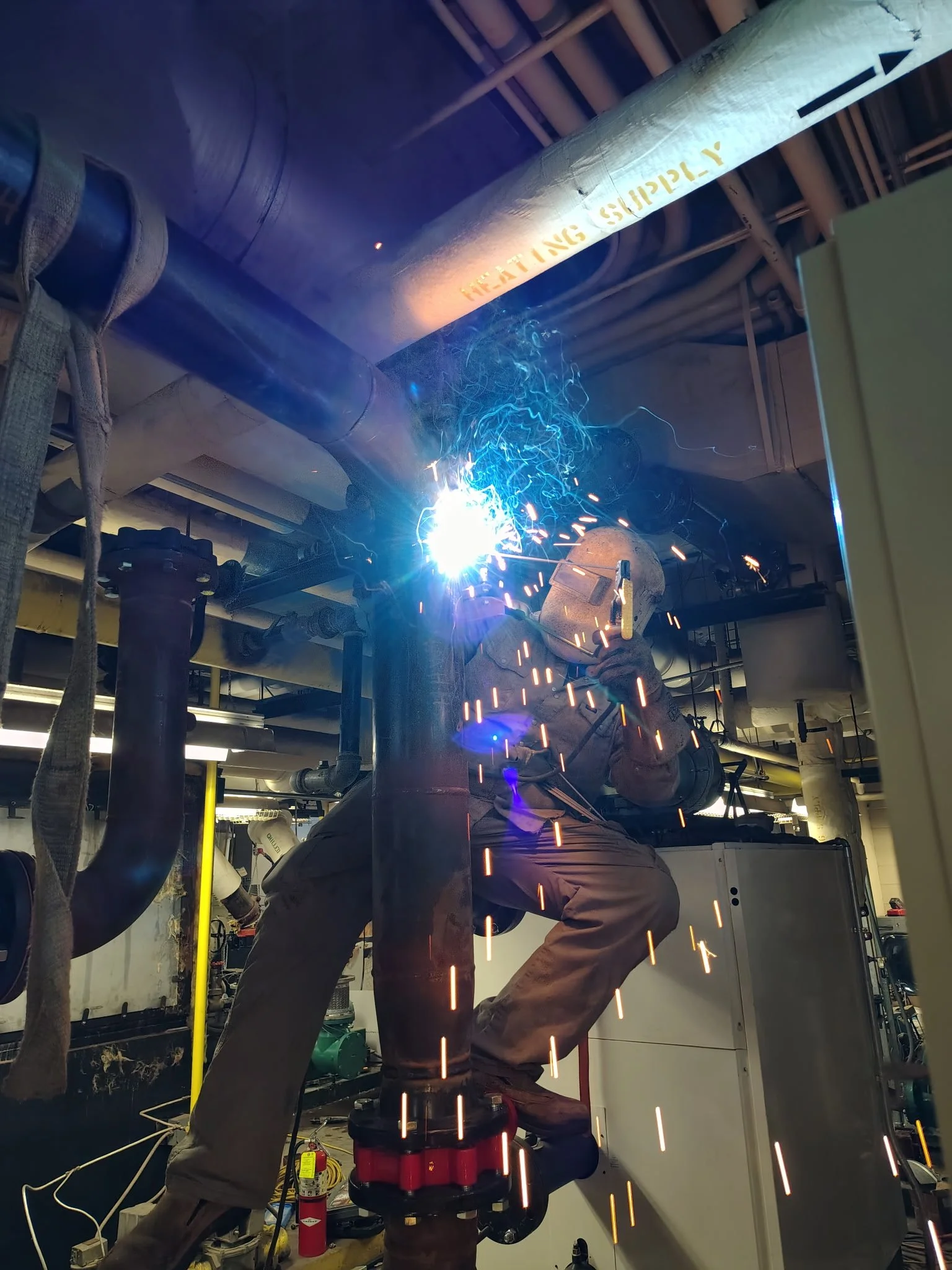 A person welding pipes in an industrial setting, with bright sparks and blue welding light visible.