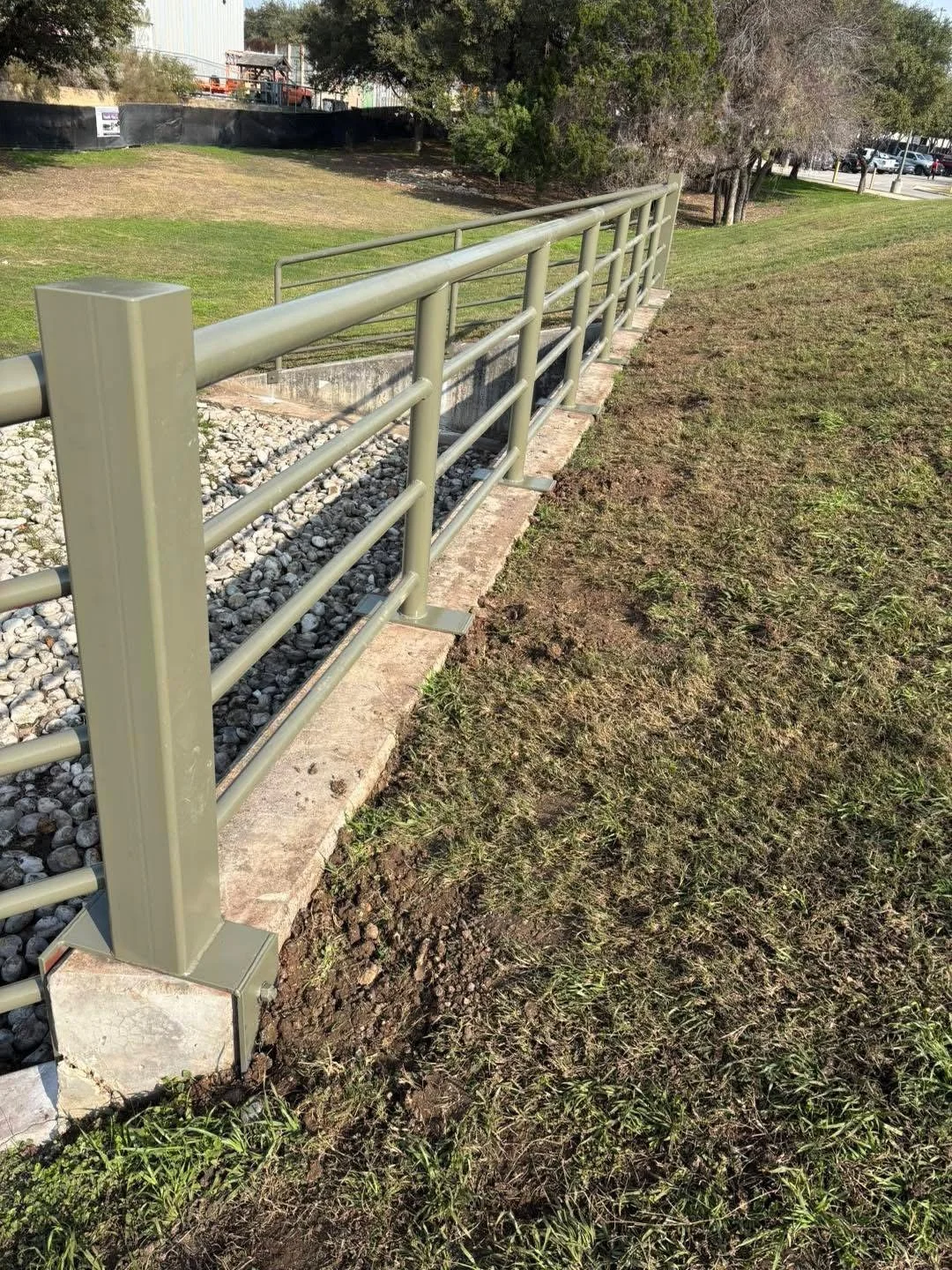 Light green metal railing along a curb on a grassy area with trees and a parking lot in the background.