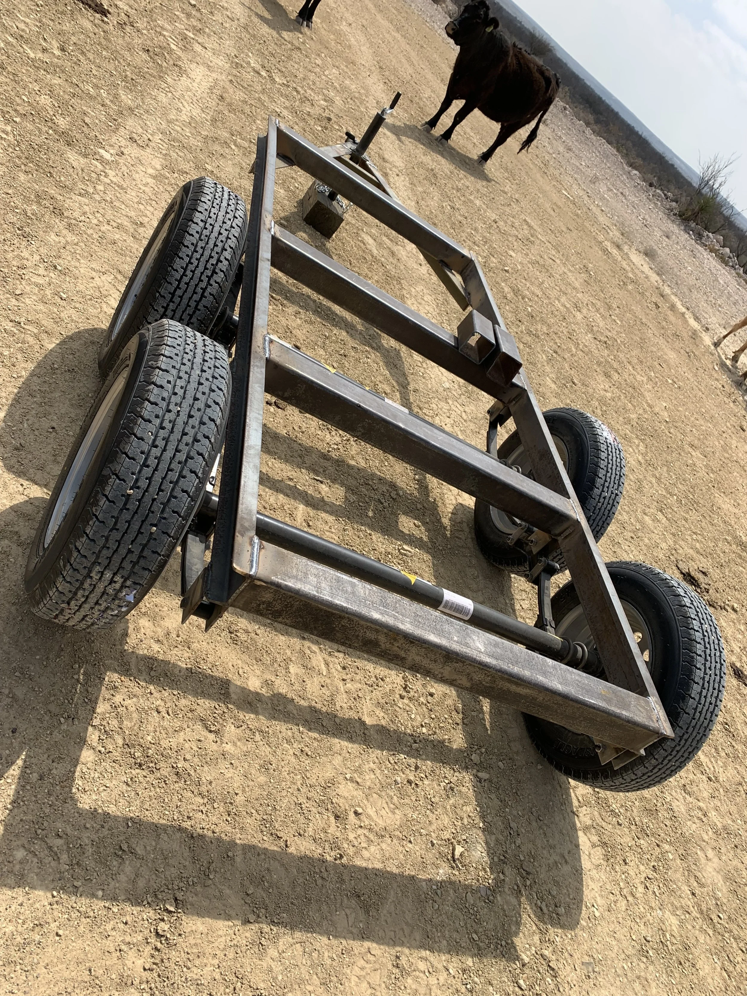 A metal cart with four black rubber tires on a dirt ground outdoors. Two horses are visible in the background, standing on the same dirt terrain under a partly cloudy sky.