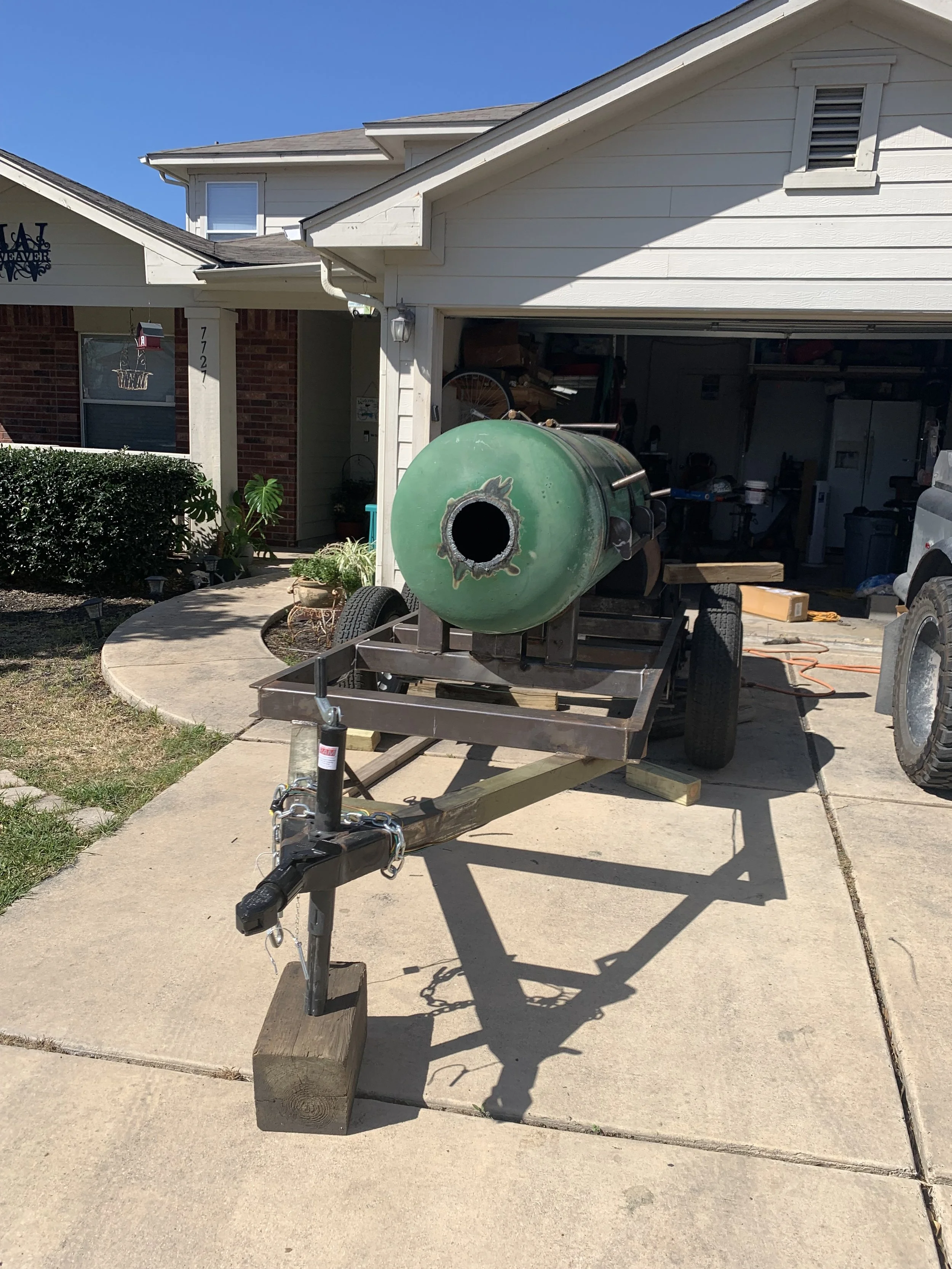 A green backyard smoker or grill on a trailer, parked in front of a house with a driveway, bushes, and various tools and objects in the garage.