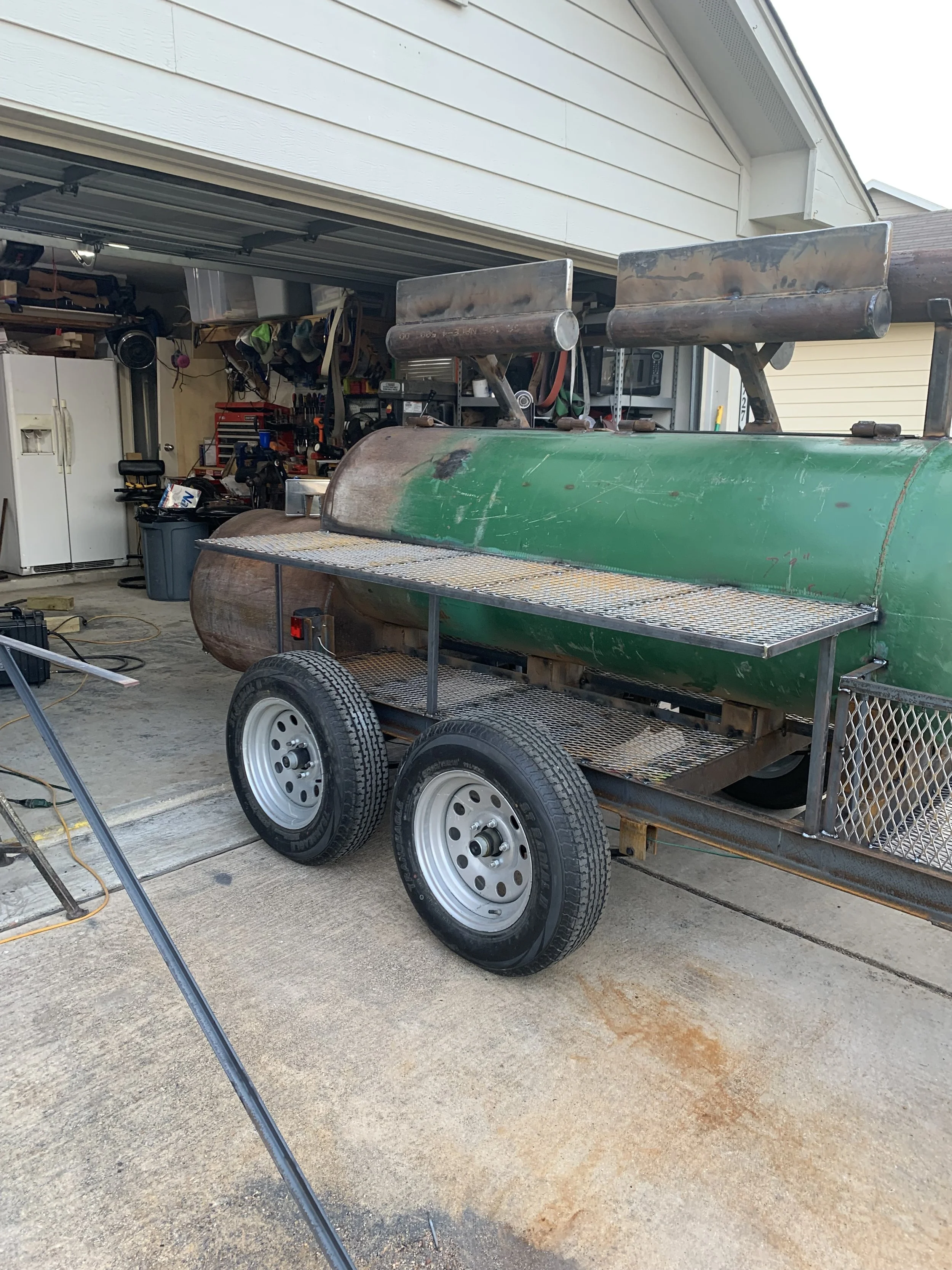 A metal trailer with green and rusty parts parked in a garage, with a fire pit or grill setup on top. The garage has tools, shelves, and a white refrigerator in the background.